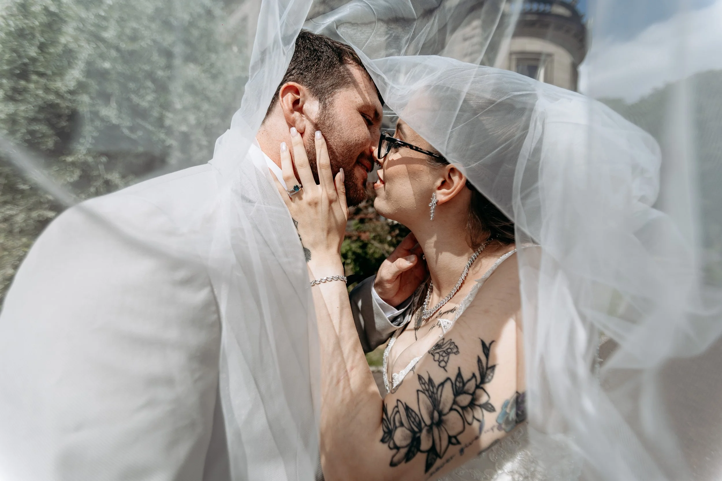 Bride and groom close-up under a veil, about to kiss outside with a historic building in the background.