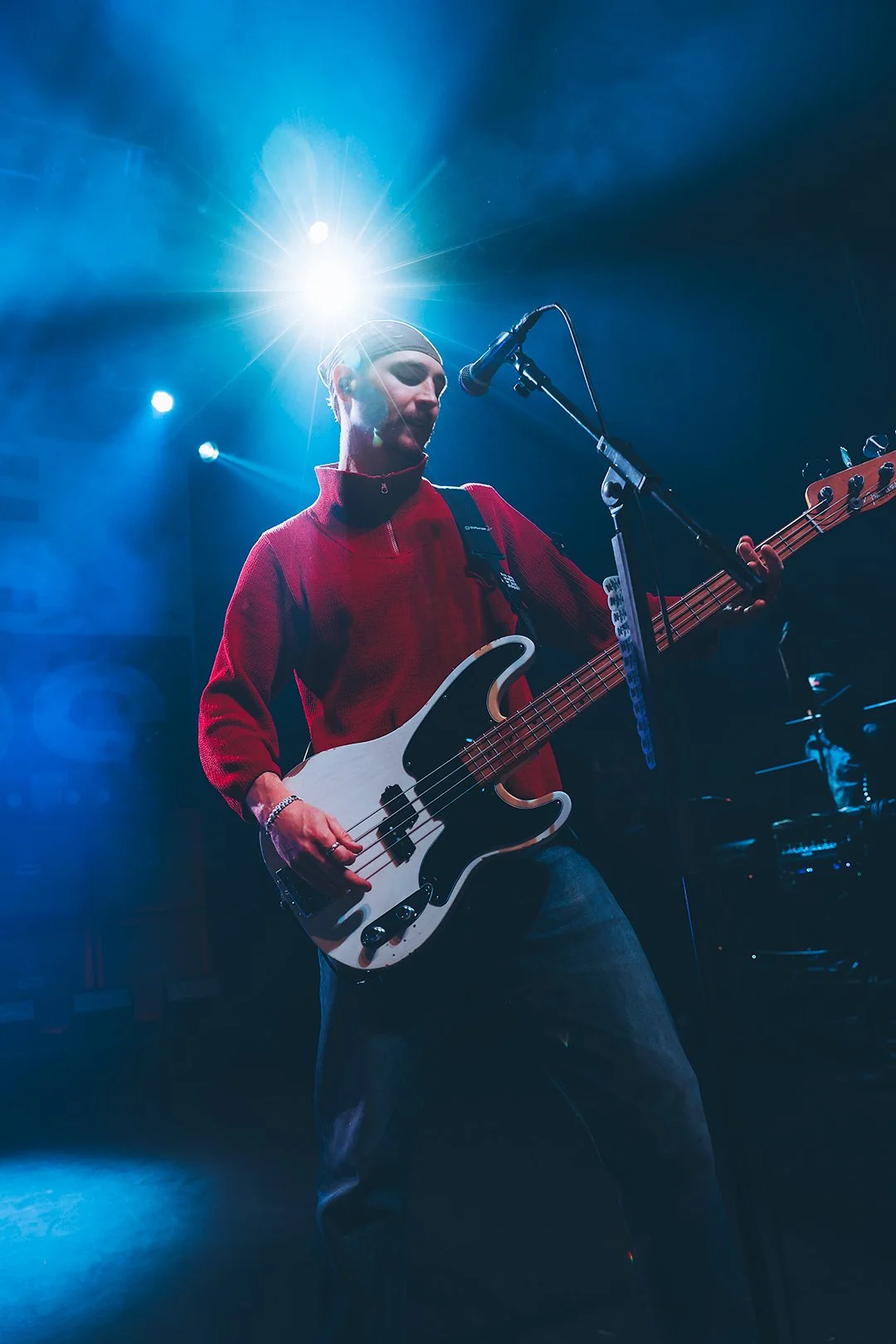 A musician wearing a red jacket playing a bass guitar on stage with blue lighting and fog.