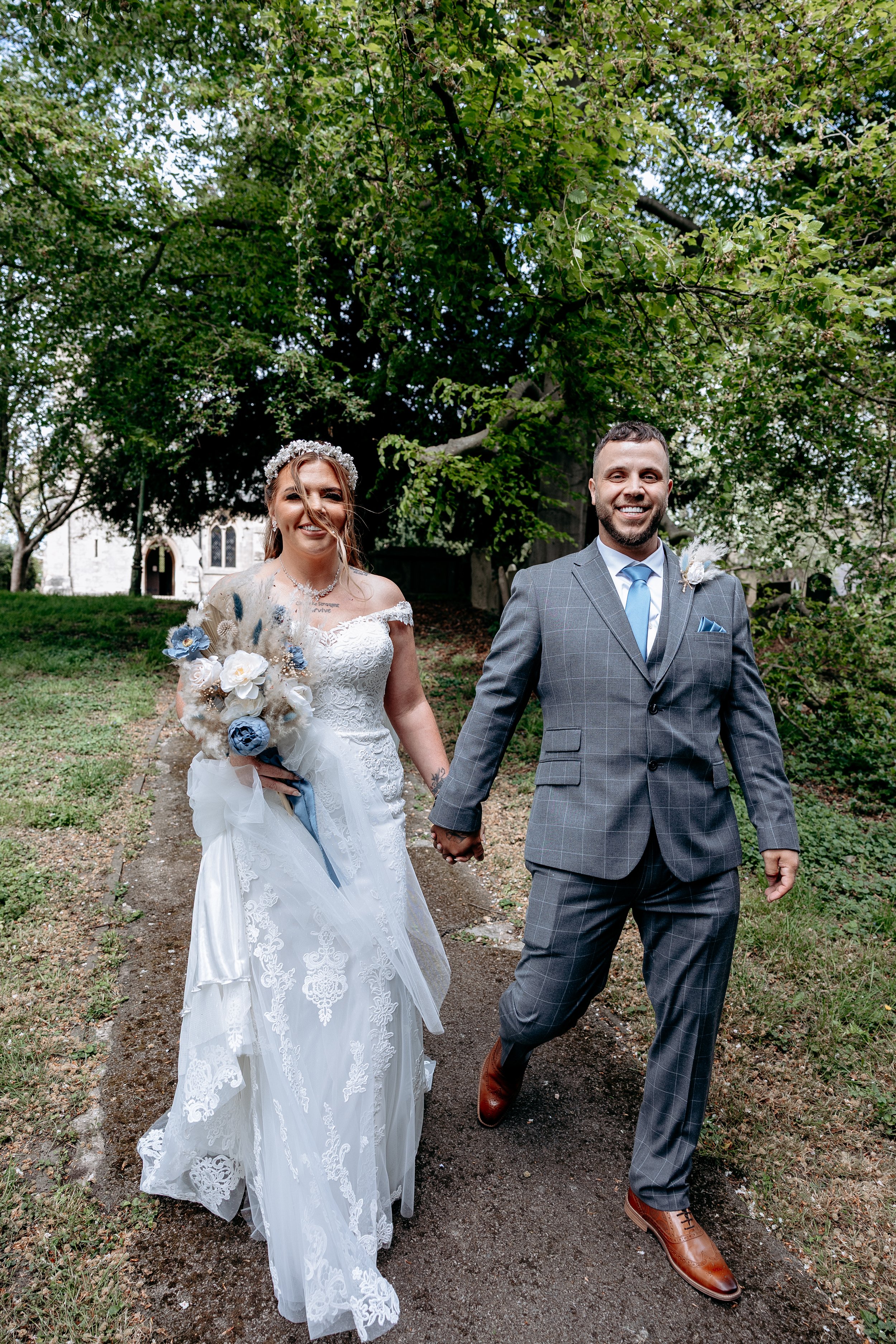 A smiling bride and groom holding hands walking outdoors on a dirt path surrounded by green trees.