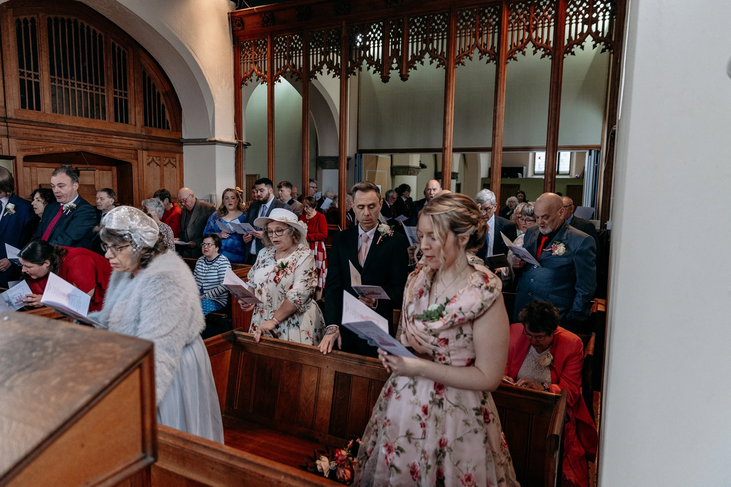 A congregation of people standing in a church, holding hymn books or programs, during a wedding or religious service. The church features wooden pews and decorative wooden paneling.