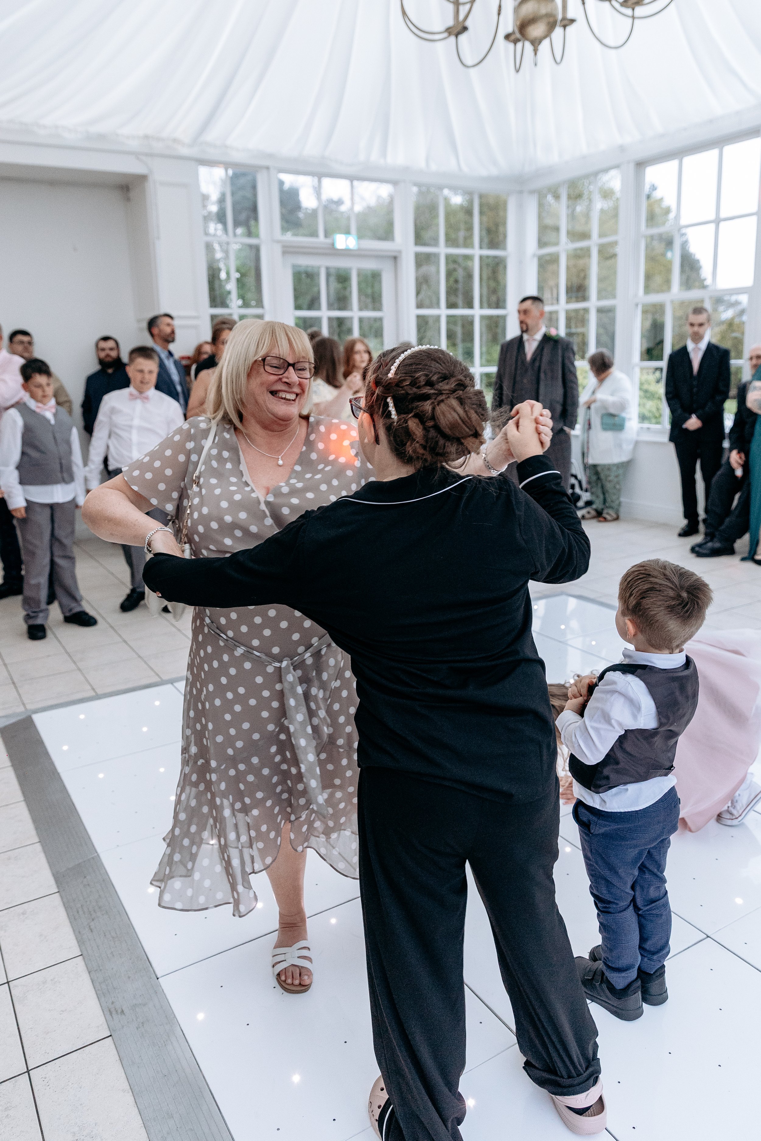 Woman dancing with young girl at a wedding reception in a bright, elegant venue with large windows and guests in the background.