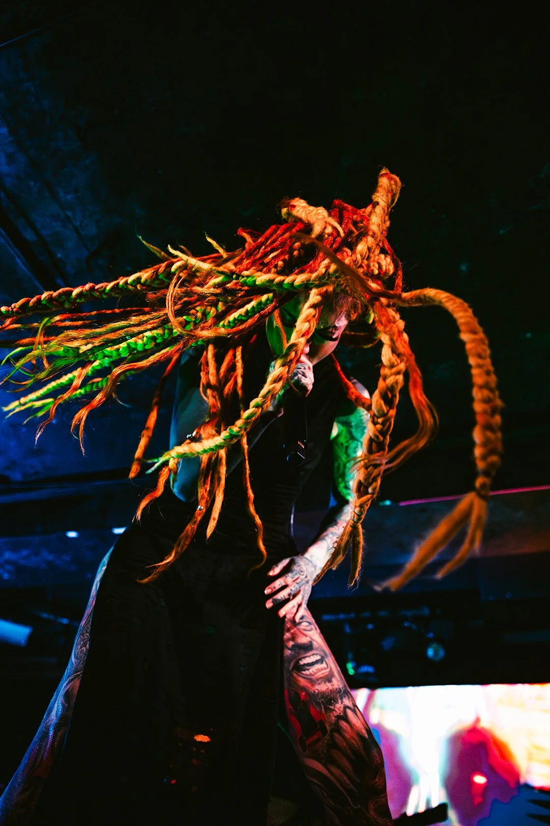 A performer with long, colorful dreadlocks and tattoos singing into a microphone on stage under colorful lights.