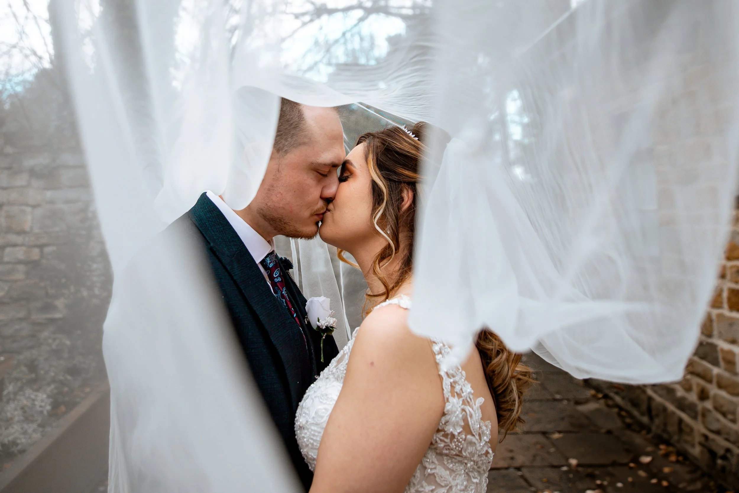 A bride and groom sharing a kiss, seen through the flowing veil on their wedding day outside near brick walls.