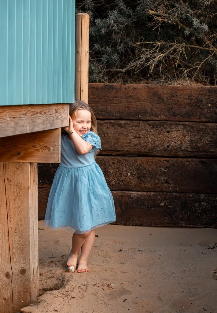 Young girl in a blue dress standing barefoot on sandy ground next to a wooden structure, smiling while leaning against a post.