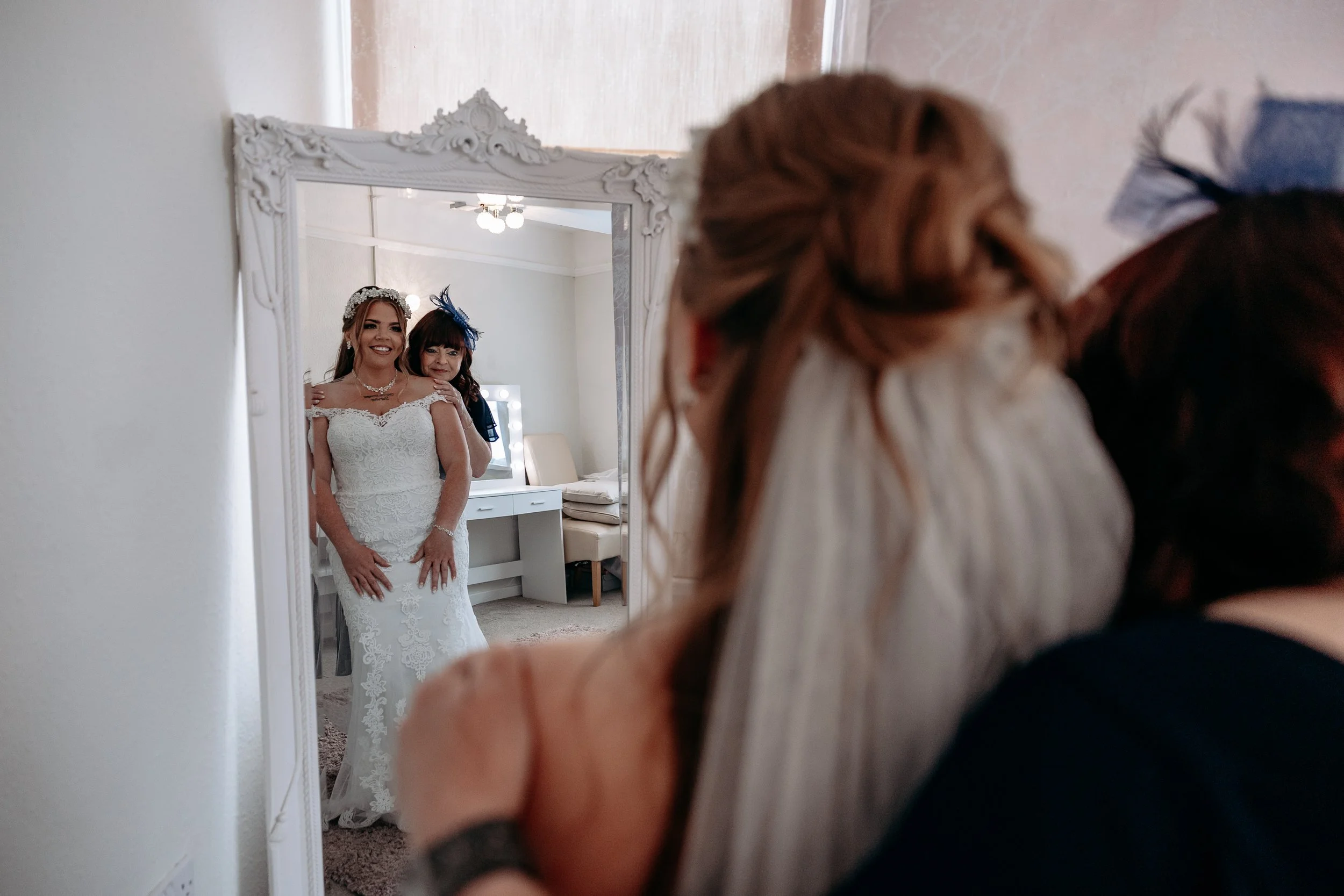 A bride in a white lace wedding dress standing in front of a large ornate mirror, being helped with her dress by a woman. Both women are smiling, and the scene is indoors with soft lighting.
