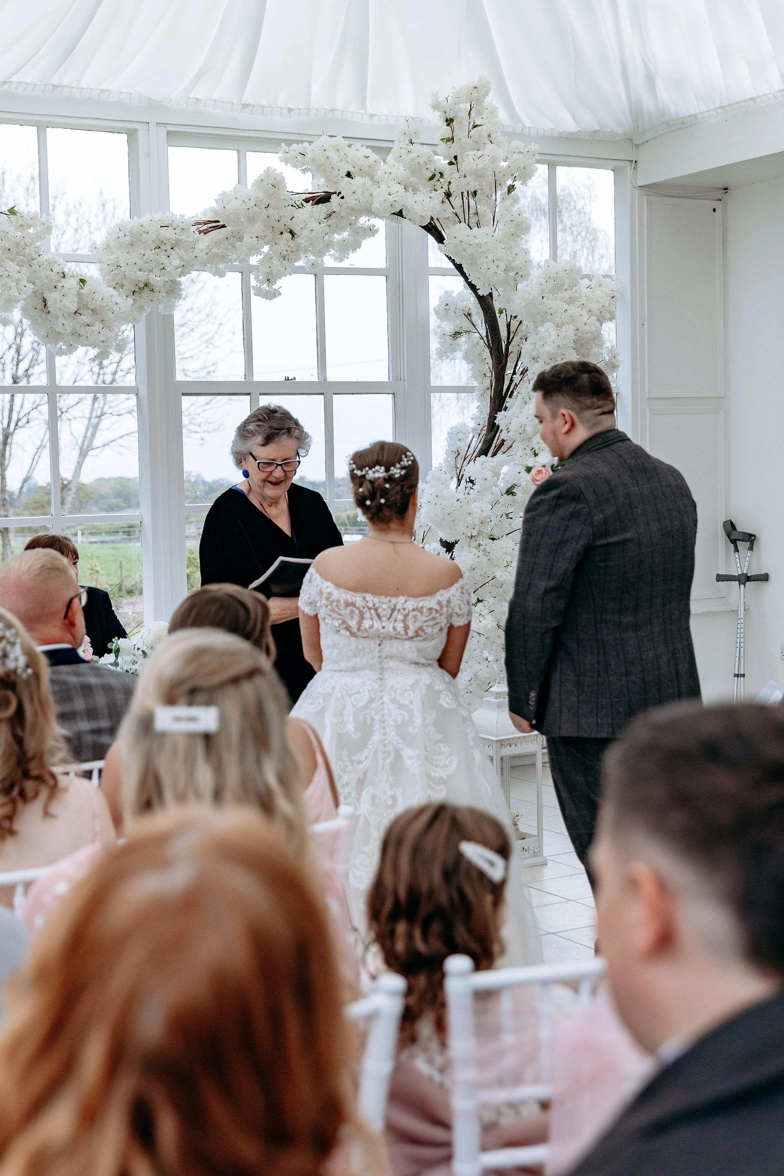 A wedding ceremony taking place indoors with a officiant, bride, and groom standing before a large floral arrangement and gathered guests.
