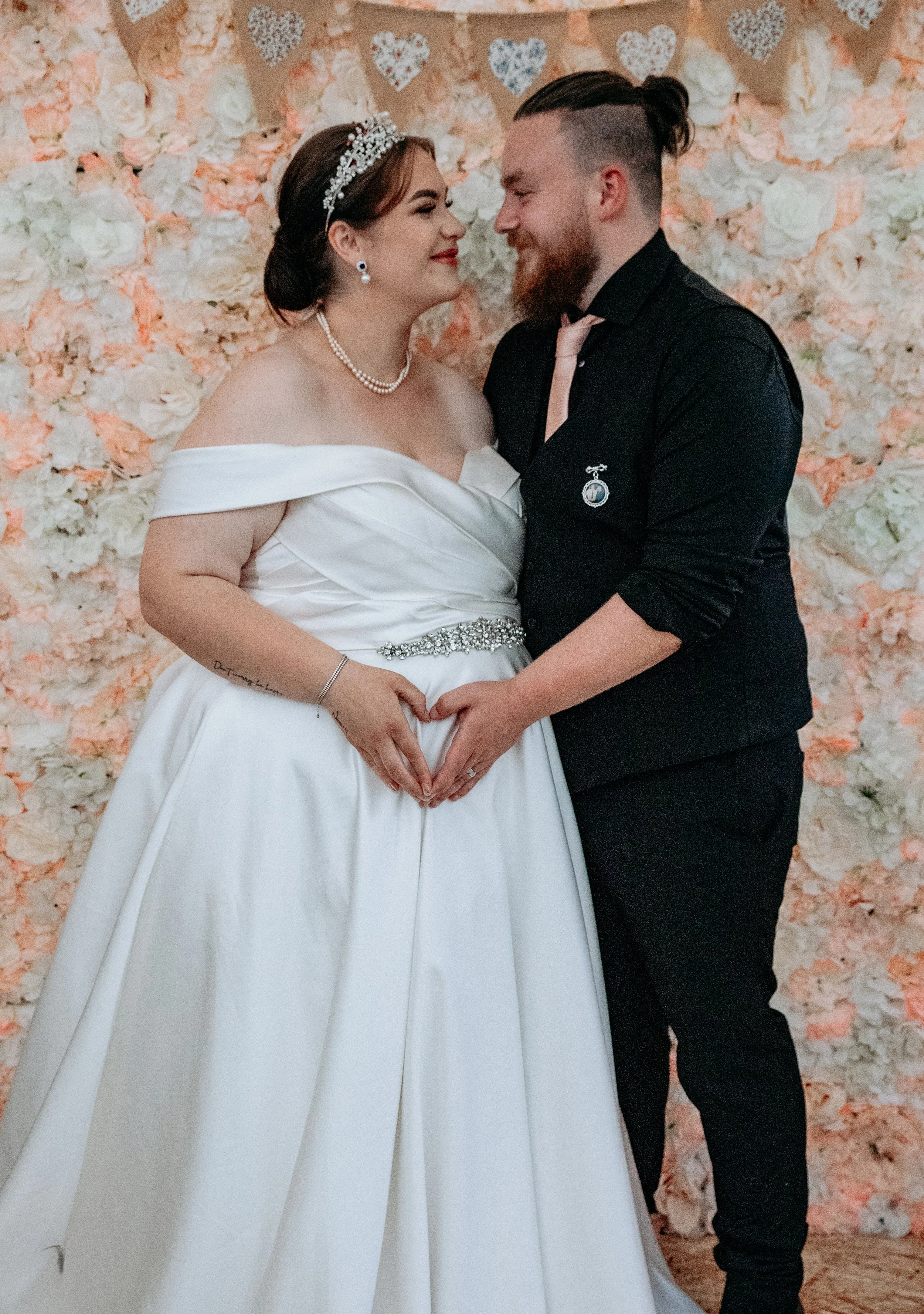 A bride and groom standing close, holding hands in front of a floral backdrop with heart decorations, smiling at each other on their wedding day.