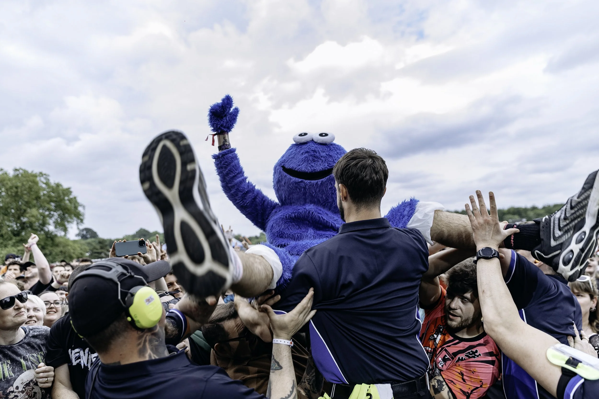 A large crowd at an outdoor event lifting a person in a blue cookie monster costume into the air. The person in costume has big eyes and a black smile, and is waving with one hand while holding an object in the other. The crowd consists of people tak