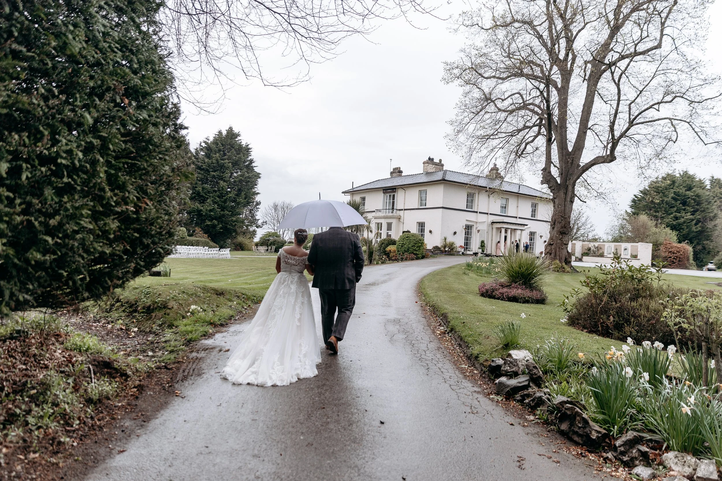 A bride in a white wedding dress and a groom in a dark suit walking on a wet driveway under an umbrella, heading towards a large white house on a rainy day.