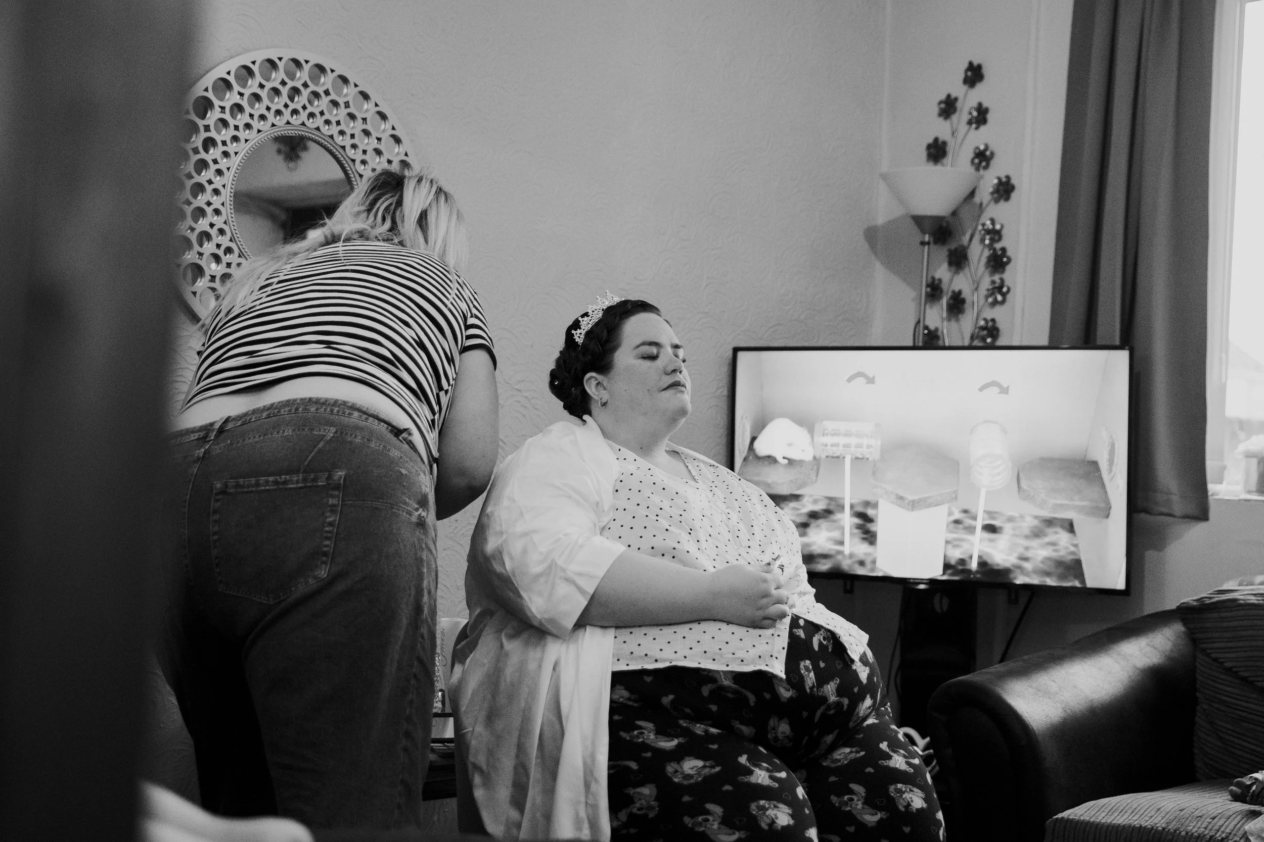 A woman wearing a crown preparing for a braid on a seated woman in a living room, with a TV showing a 3D model of food, and a wall with a mirror, lamp, and decorative metal wall art.