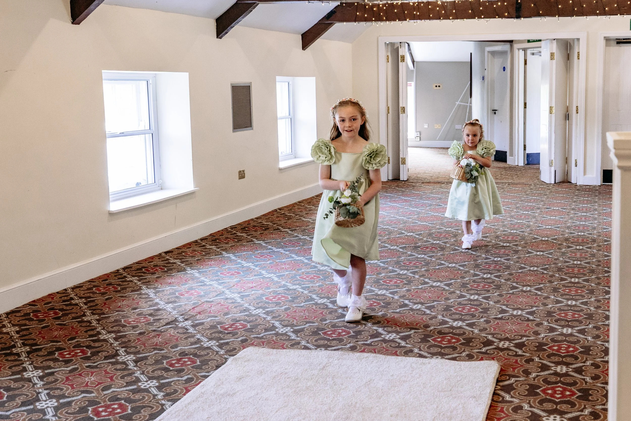Two young girls in light green dresses with puffed sleeves walking down a patterned carpeted hallway, each carrying a small basket of flowers, in a well-lit indoor setting with white walls and windows.