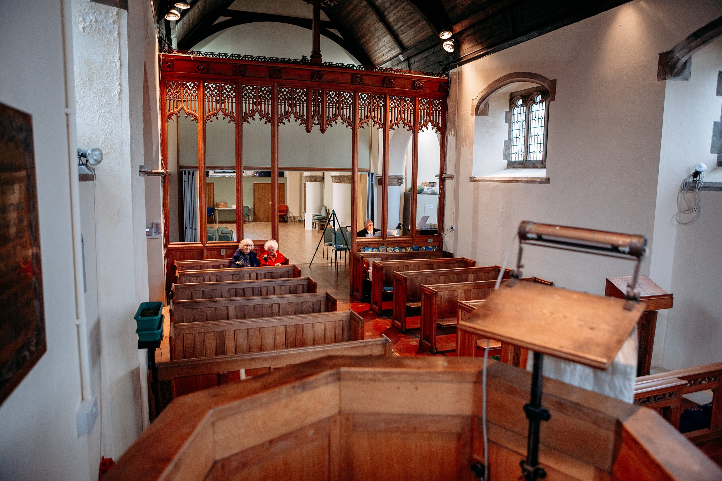 Interior of a church with wooden pews, three elderly women sitting, and a choir stall at the front. The view is from the pulpit, showing the sanctuary and stained glass windows.