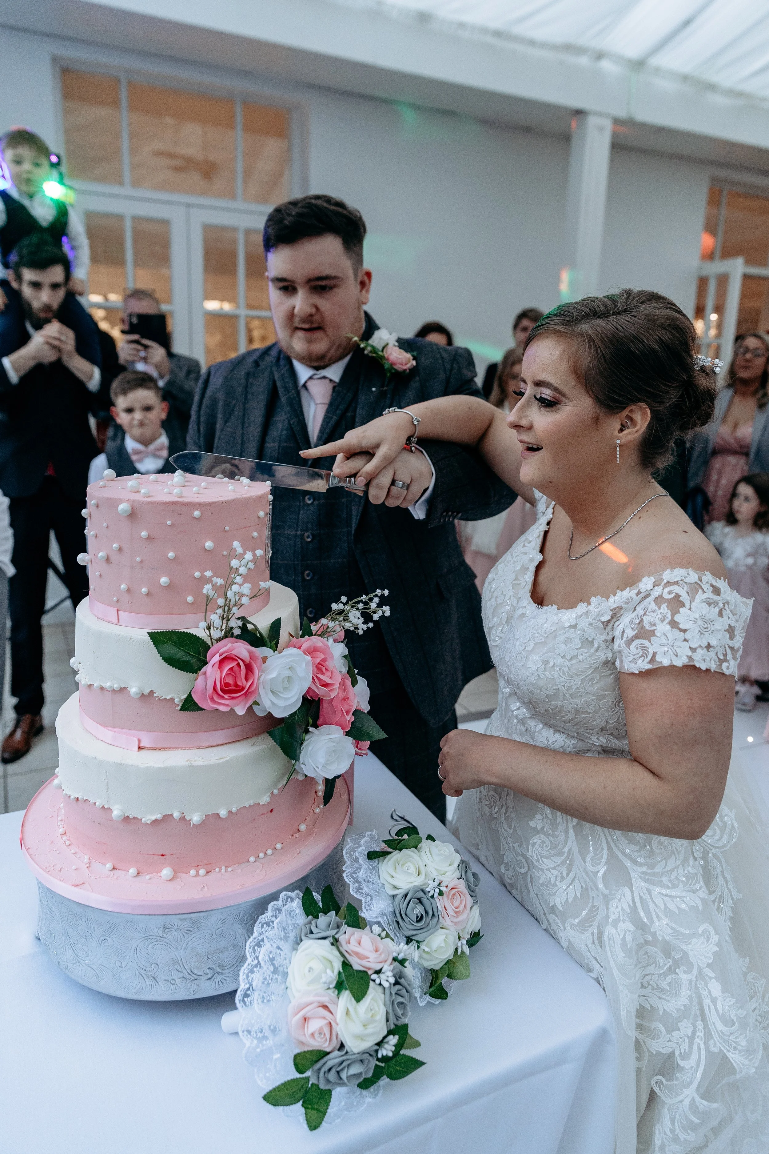 Bride and groom cutting a three-tier pink and white wedding cake decorated with flowers, with guests watching in the background.
