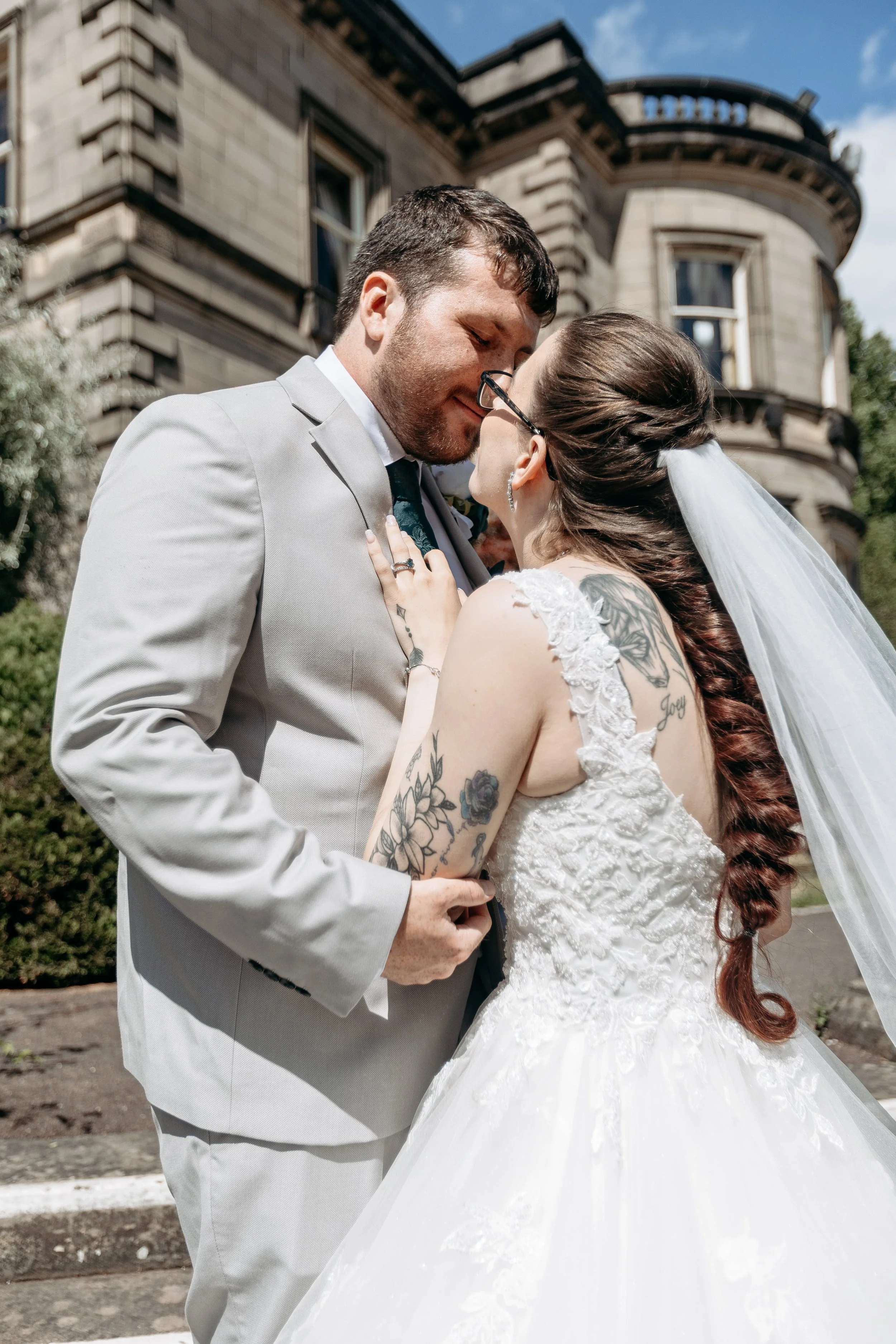 A bride and groom share a kiss outdoors in front of a historic building on a sunny day.