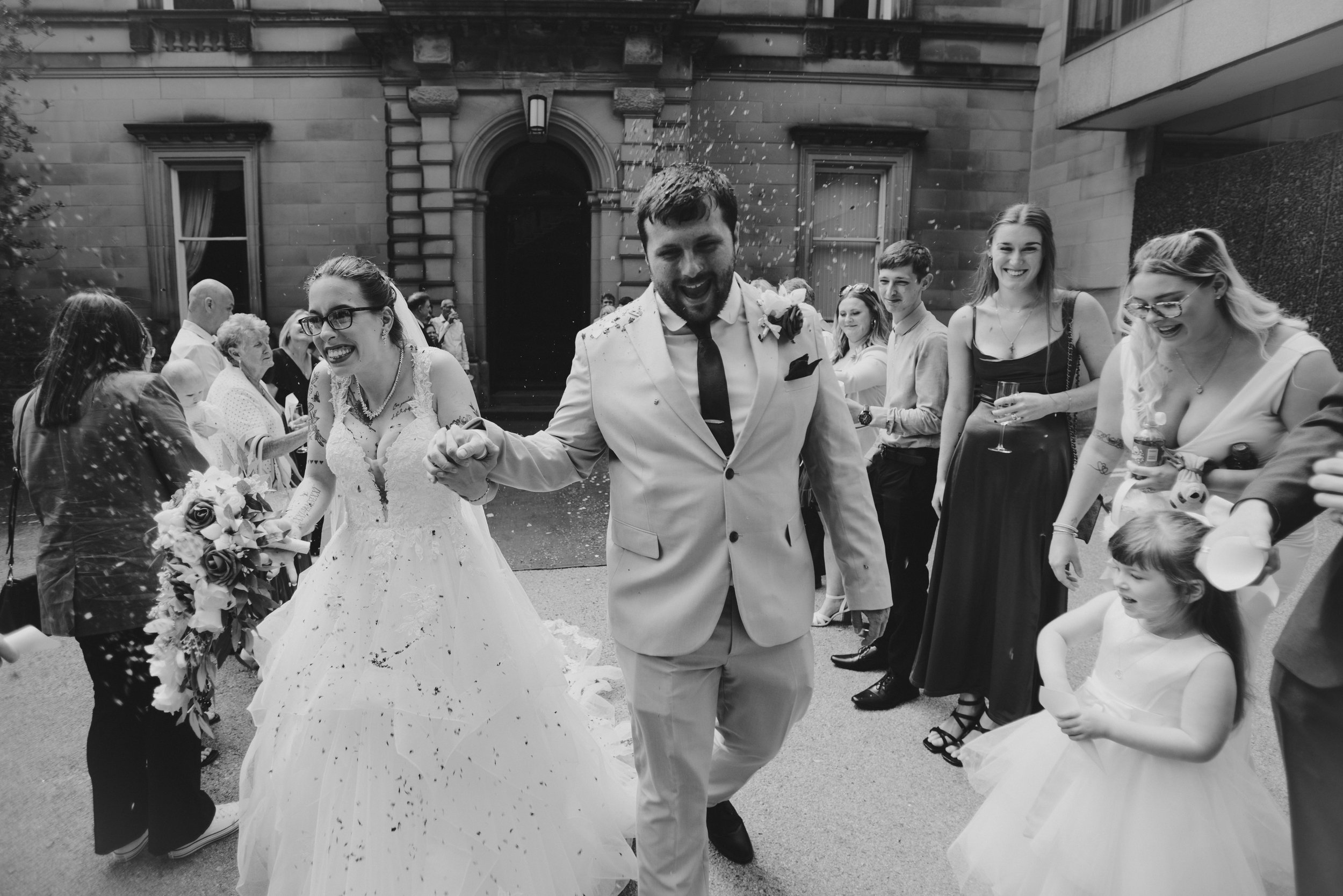 A black and white photo of a wedding celebration showing a bride and groom smiling and holding hands as guests throw confetti. The bride wears a lace wedding dress and holds a bouquet, and the groom wears a light-colored suit with a tie and boutonniè
