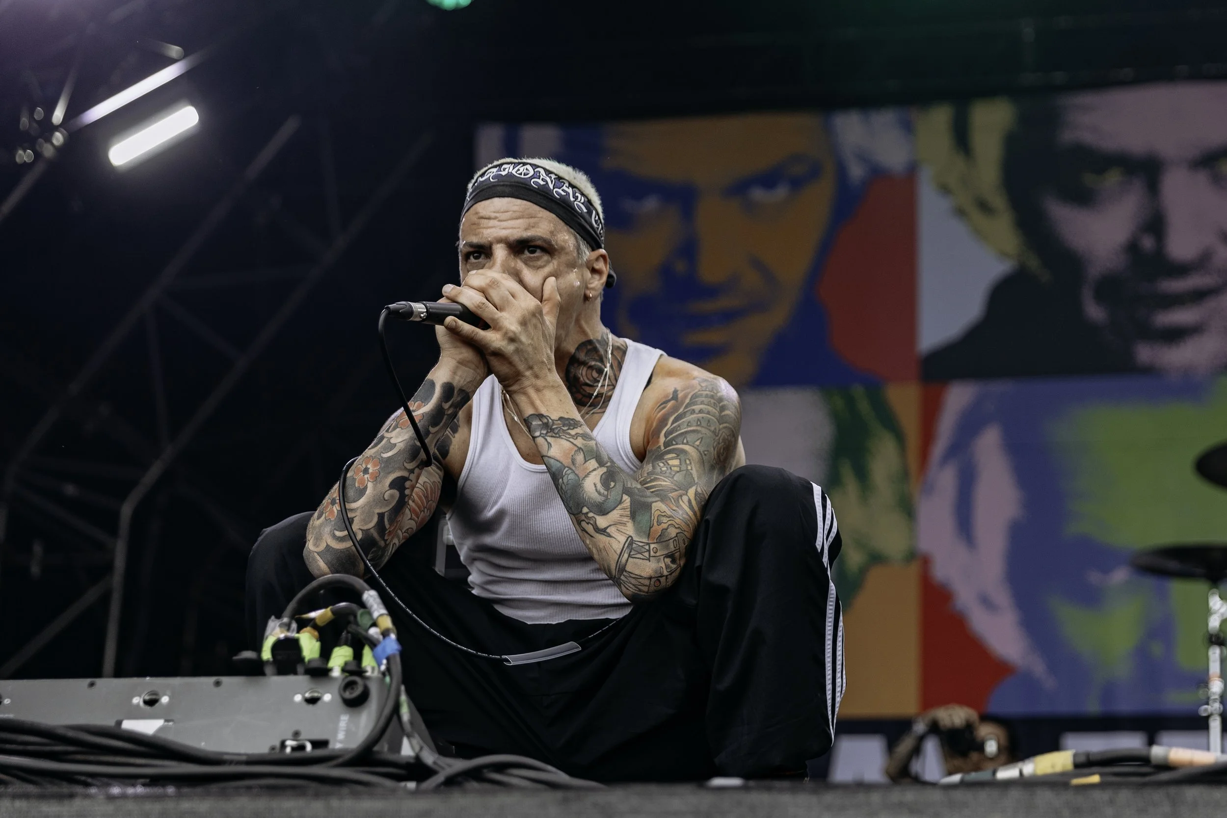 A musician with tattoos and wearing a white tank top and a headband, crouching on stage and holding a harmonica to his mouth, with a microphone in hand. Behind him are colorful pop art portraits on a large screen.