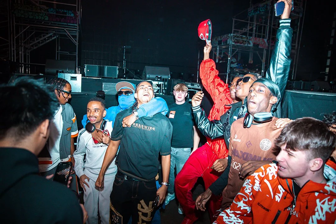 Group of young people in colorful streetwear celebrating at a lively gathering or event, with one person raising a red cap in the air. Stage equipment is visible in the background.