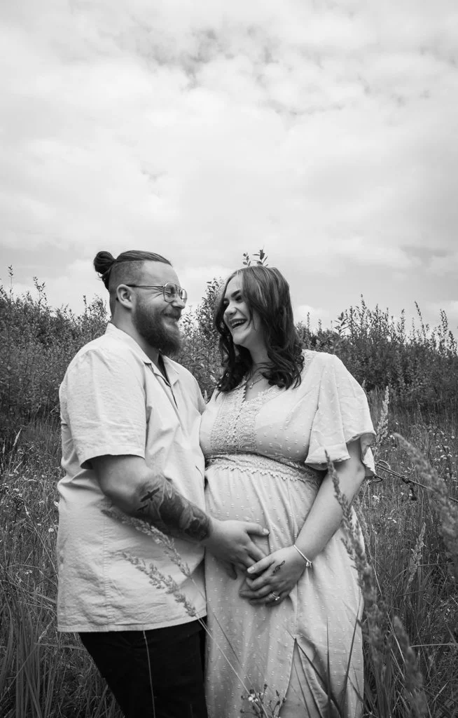 A pregnant woman and a man standing in a field, holding hands and smiling at each other.
