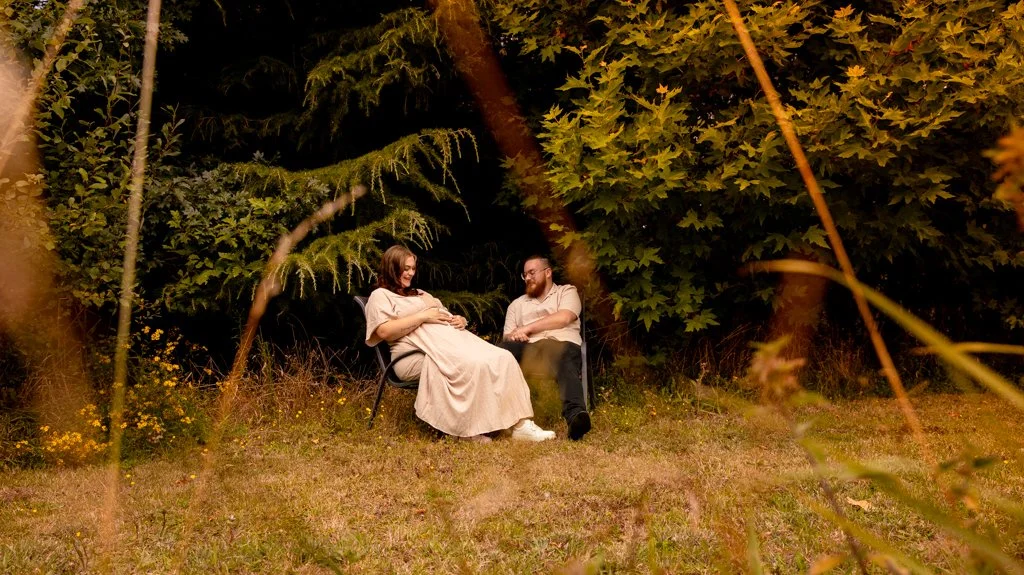 A pregnant woman and a man sitting on chairs outdoors in a wooded area at night, smiling and looking at each other.
