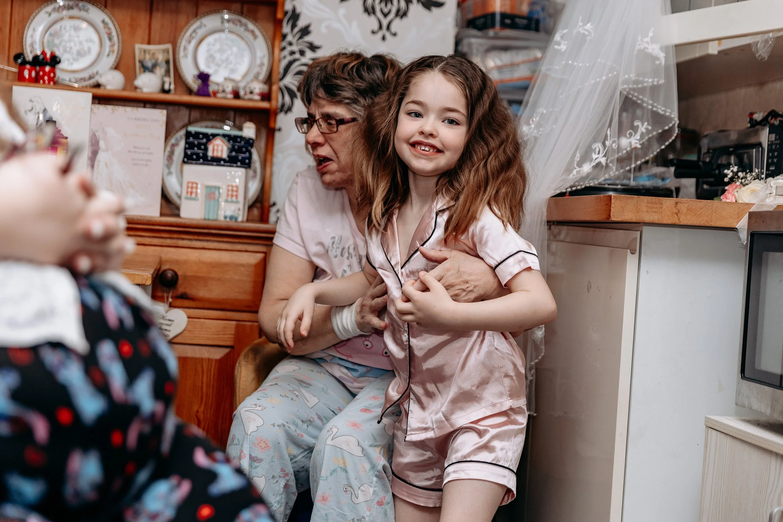 A young girl in pink satin pajamas smiling as an older woman holds her from behind in a cozy kitchen with decorative plates and photos on the shelves.