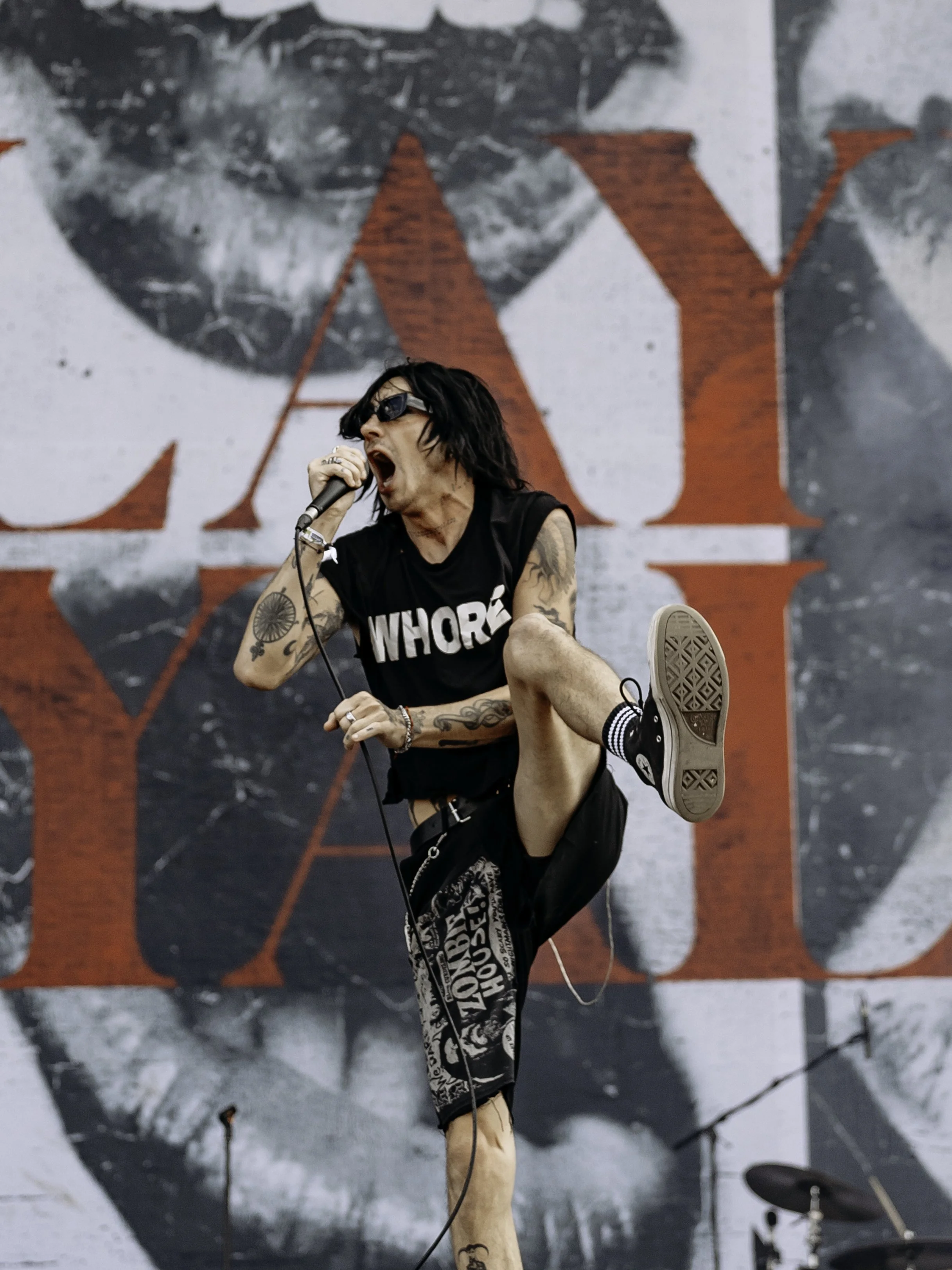 A male singer with black hair, sunglasses, and tattoos, wearing a black sleeveless shirt and shorts, performing on stage with a microphone, with a large backdrop featuring a black and white image and red letters.