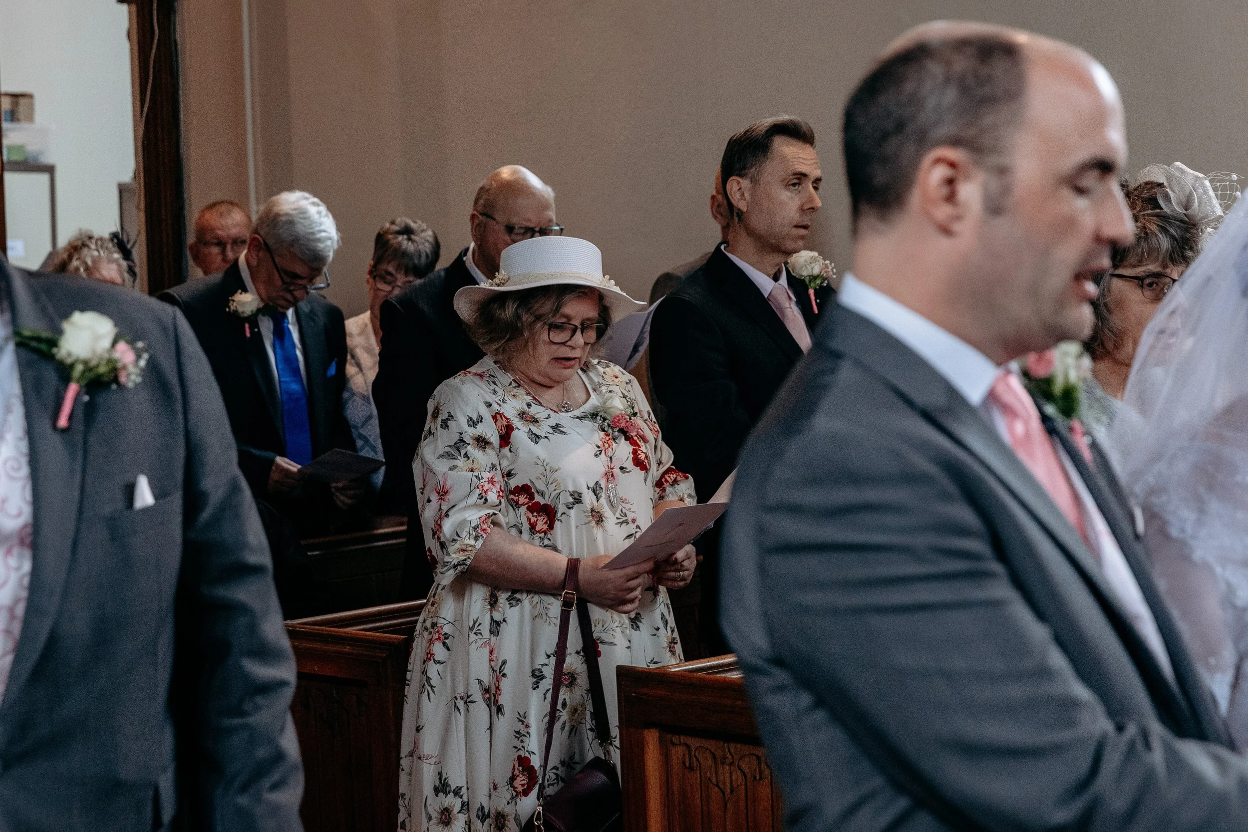A group of people attending a wedding ceremony in a church, with some reading from programs or hymnals. The focus is on a woman in a floral dress and wide-brimmed hat, and a man in a suit with a pink tie. Other attendees are visible in the background