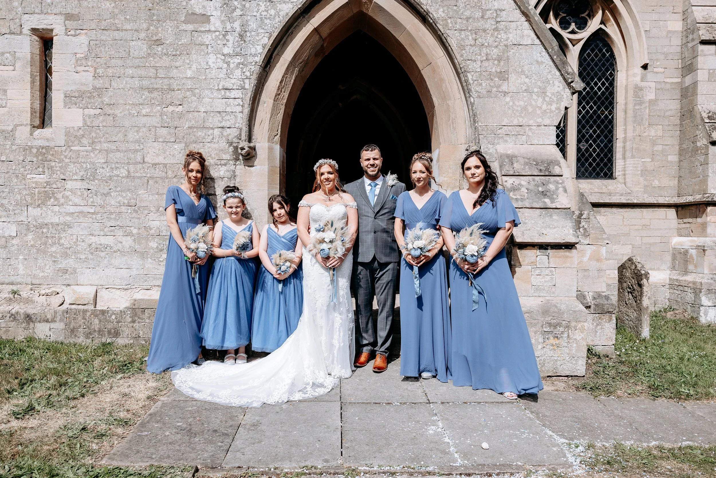 A wedding party standing outside a stone church, consisting of six women in blue dresses, a bride in a white wedding gown, and a groom in a gray suit. The women are holding bouquets and the bride is holding a bouquet.