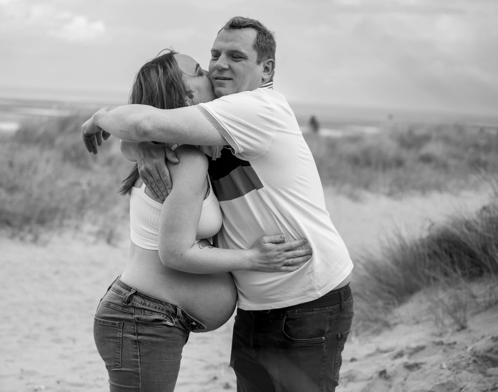 A pregnant woman and a man embracing on a beach, with the woman kissing the man's cheek, in black and white.