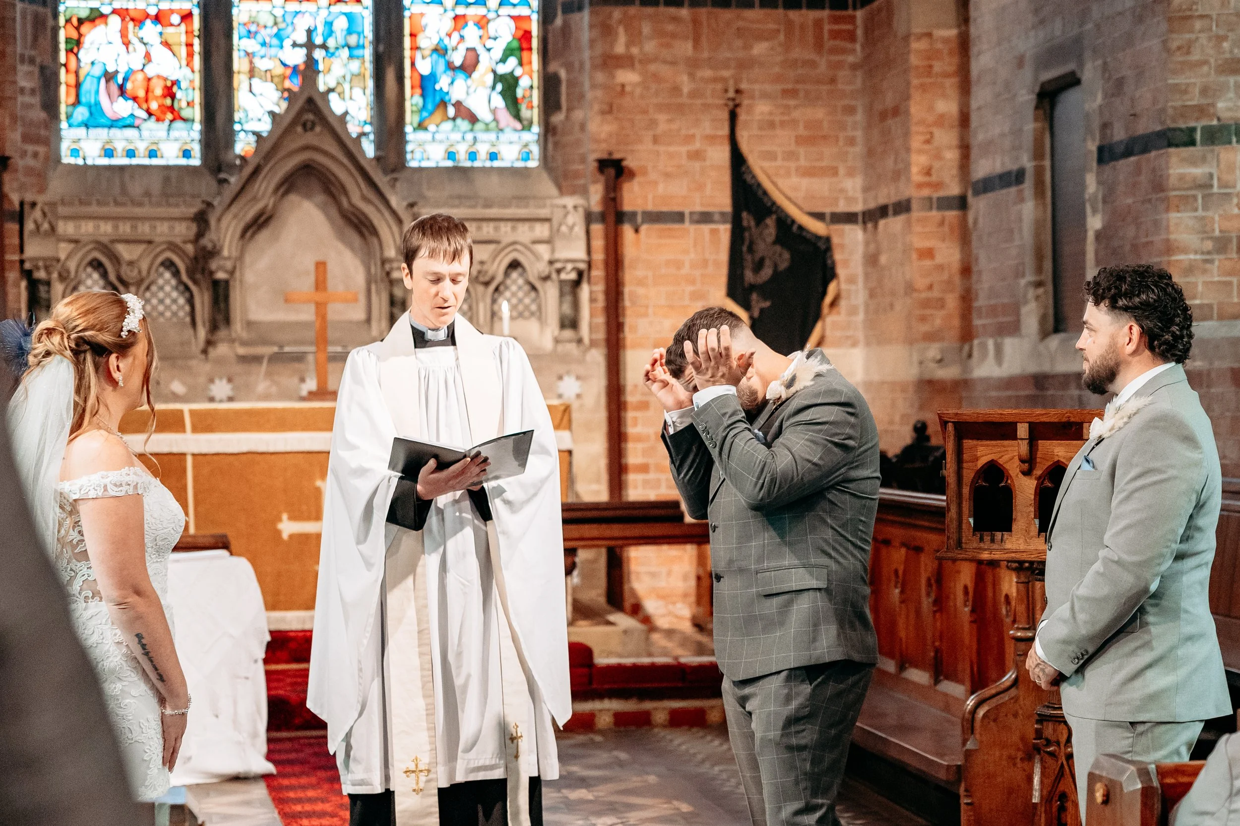 A couple gets married in a church during their wedding ceremony, with a priest officiating. The bride is wearing a white wedding dress and the groom is in a gray checkered suit. The officiant is holding a book, and there is a stained glass window and