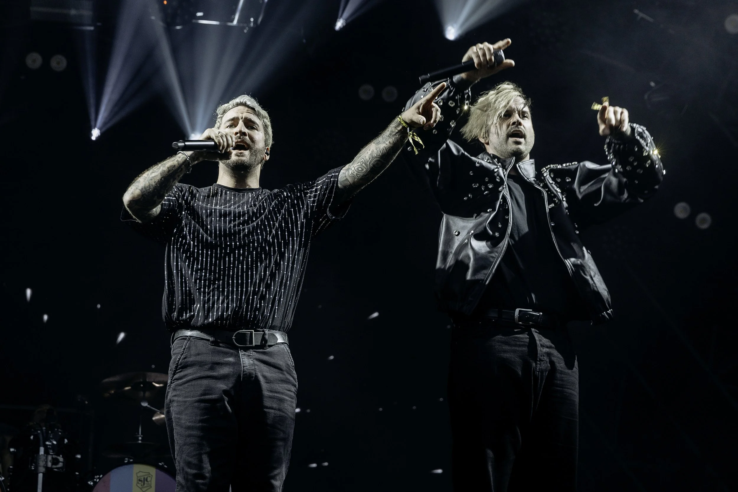 Two male performers singing on stage under bright lights, one with a microphone, both wearing dark clothing, with a drum set in the background.