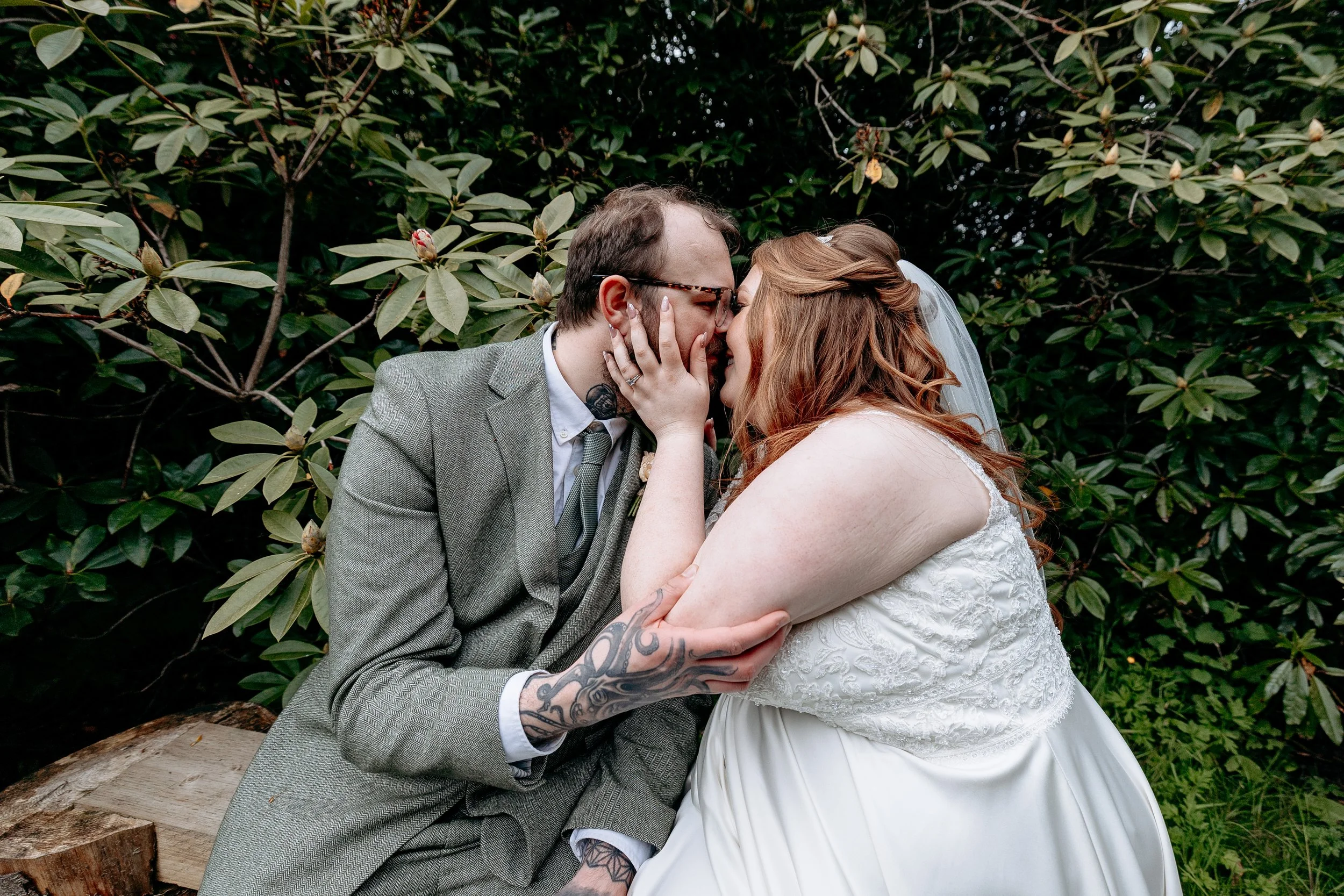 A couple sitting outdoors on a wooden bench, embracing and about to kiss, surrounded by greenery.