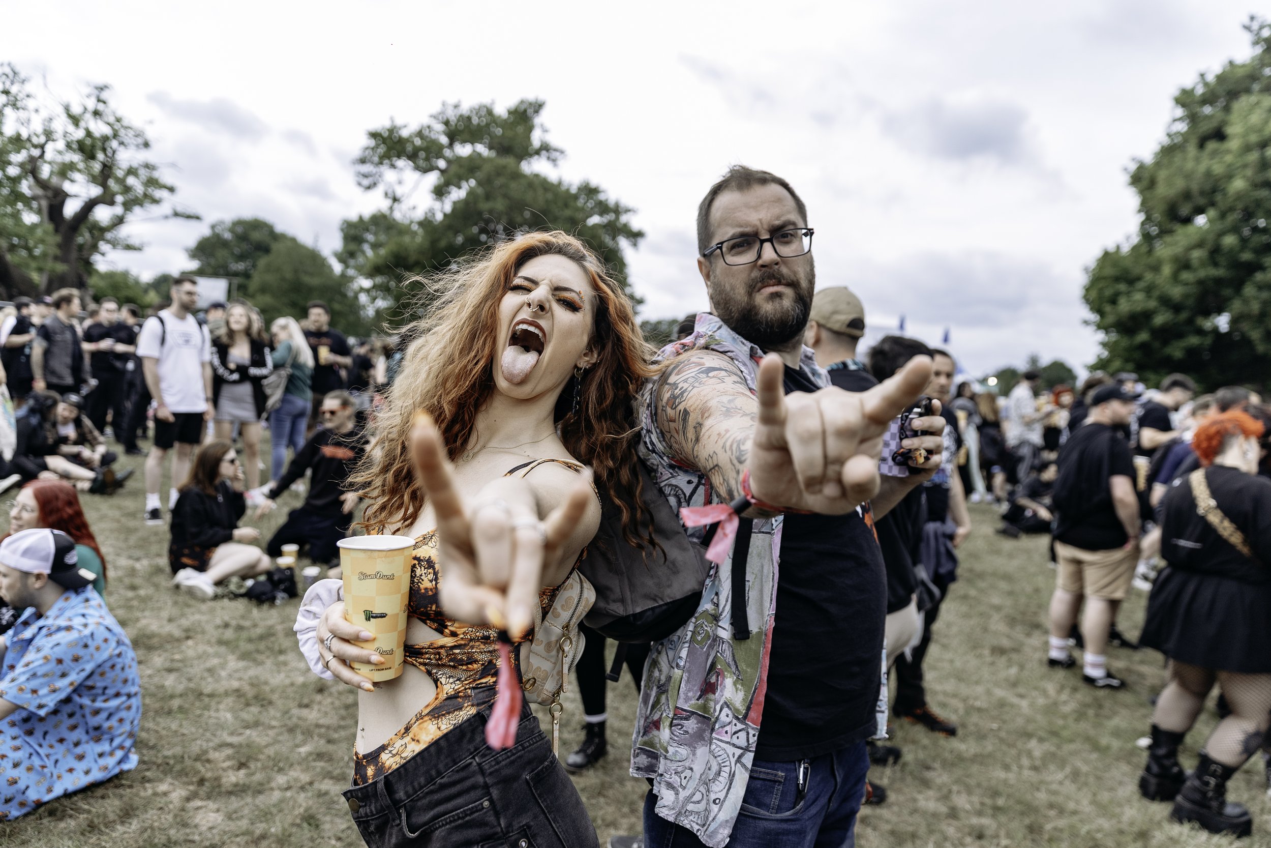 Two people in a crowd at an outdoor event. The woman on the left has red hair, is making a face with her tongue out, and is holding a cup. The man on the right has glasses and tattoos, and is pointing towards the camera with a serious expression. Man