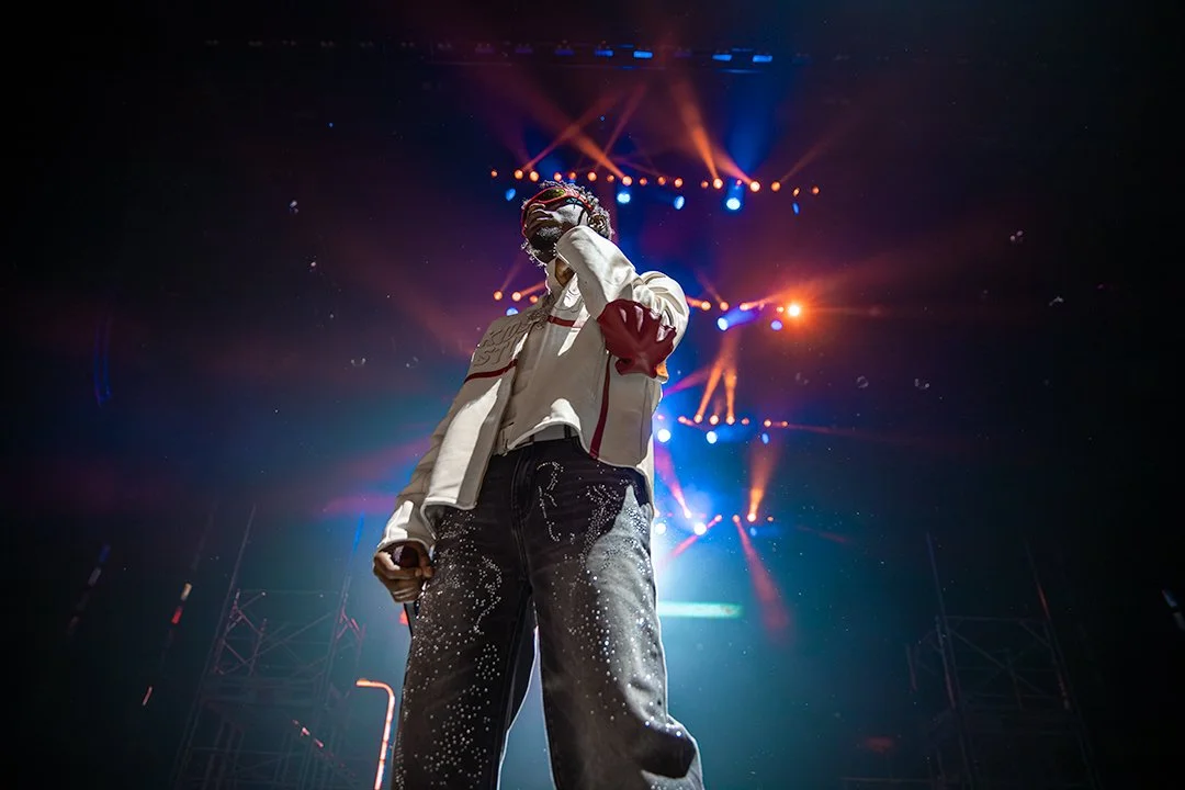Performer on stage with colorful lights shining down, wearing a cream jacket with red accents and black jeans with white paint splatters.