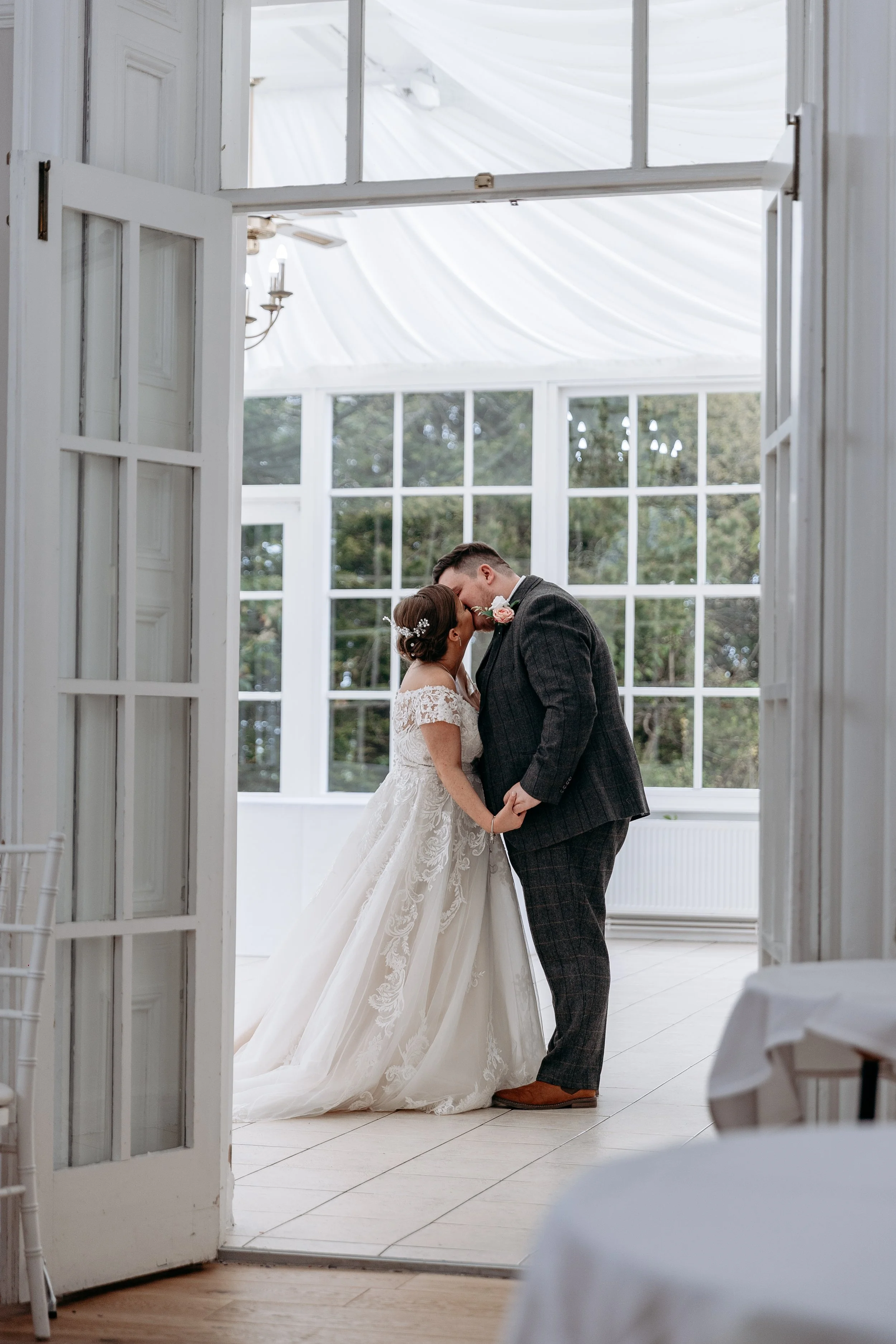 A bride and groom sharing a kiss at their wedding venue, framed by open double doors with a white interior and large window behind them showing green trees.