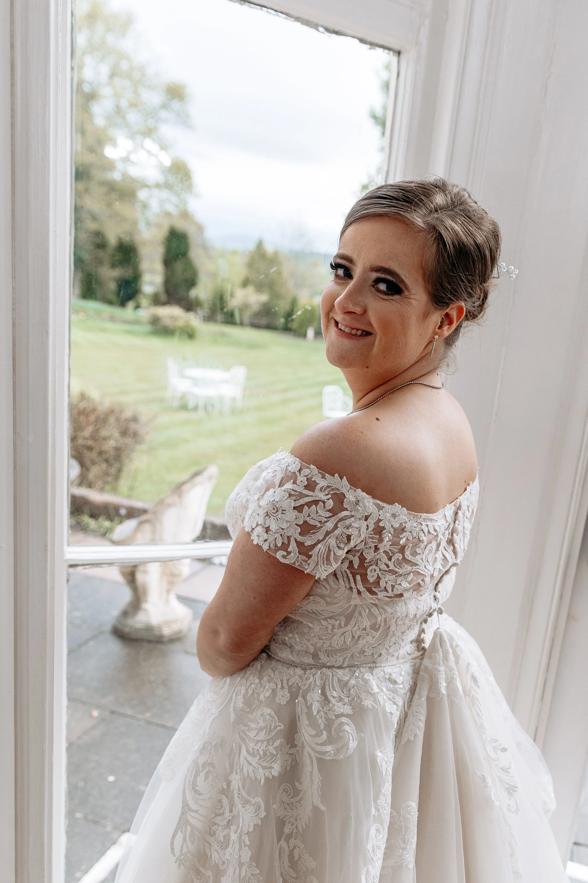 A bride in an off-shoulder lace wedding dress standing in front of a window, smiling and looking over her shoulder.