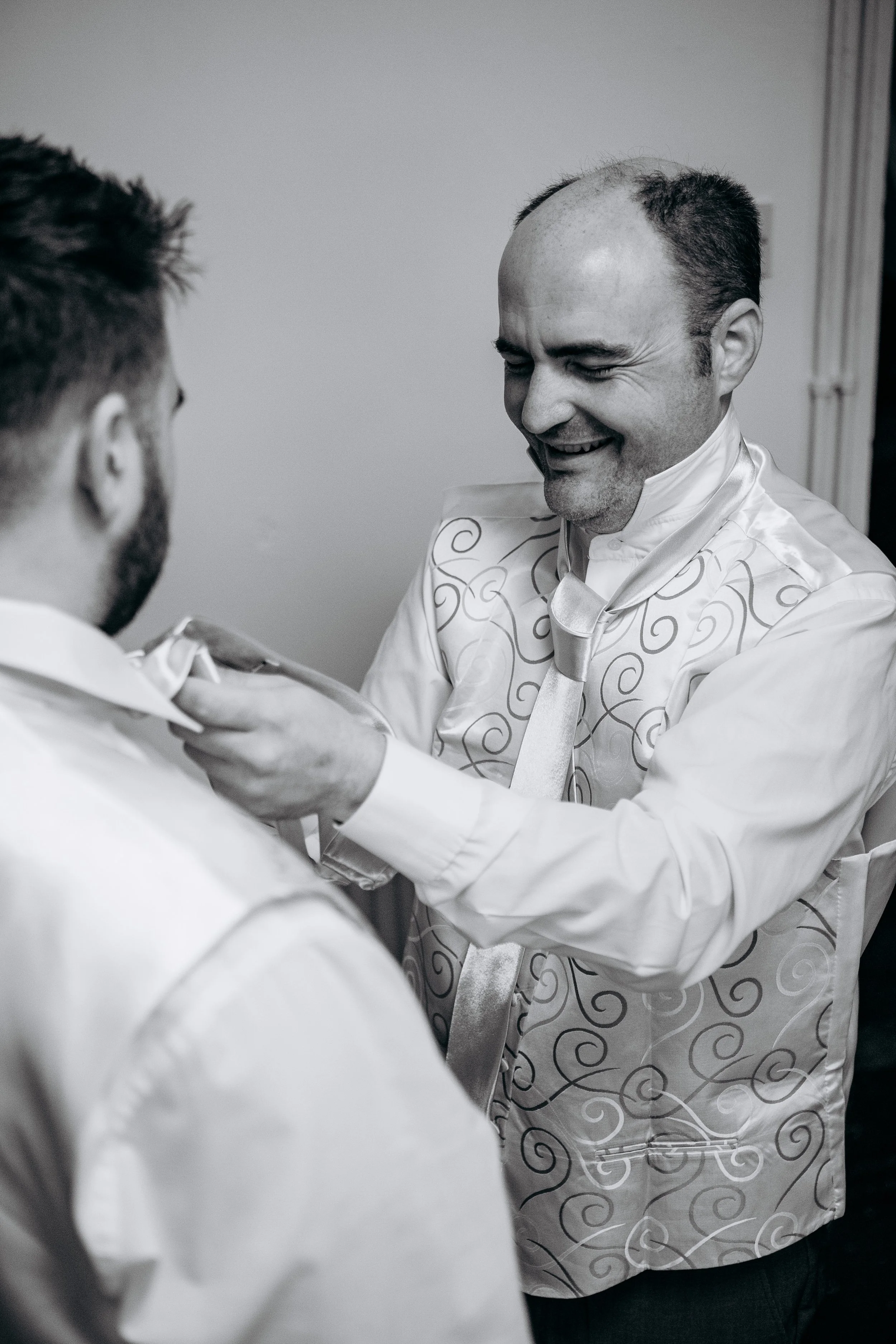 A man helping another man with his bow tie, both dressed in formal attire, smiling in a candid moment.