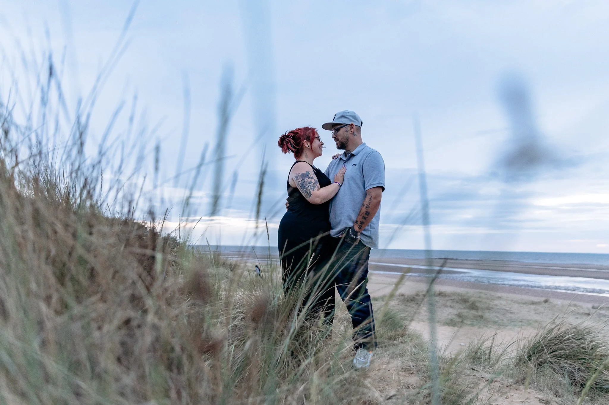 A couple standing close together on a sandy beach, gazing into each other's eyes during sunset, with tall grass in the foreground and the ocean in the background.