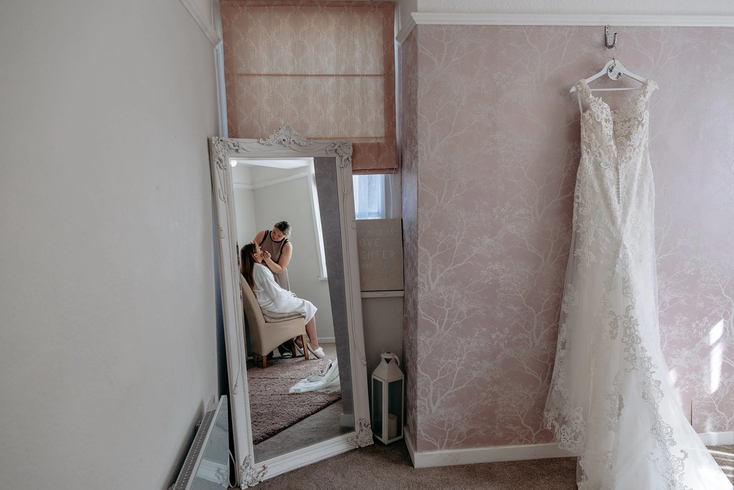 A wedding dress hanging on a wall with a mirror reflecting a woman sitting in a chair having her makeup done, in a softly lit room with pink patterned wallpaper.