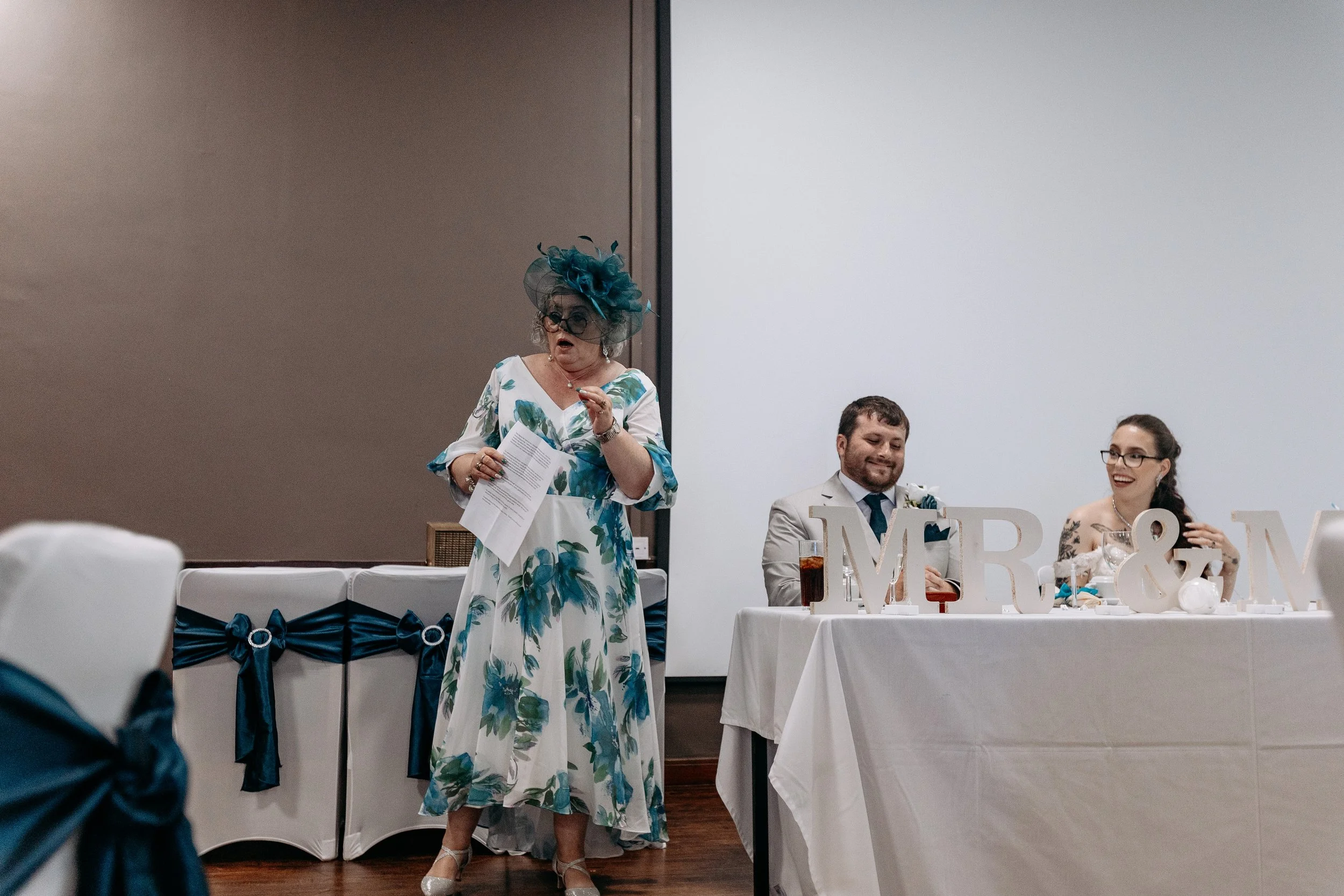 A woman giving a speech at a wedding reception, standing beside a table with a bride and groom seated. The woman is dressed in a floral dress and large hat, holding papers. The groom is smiling, and the bride is laughing. Decorative 'MR & MRS' letter