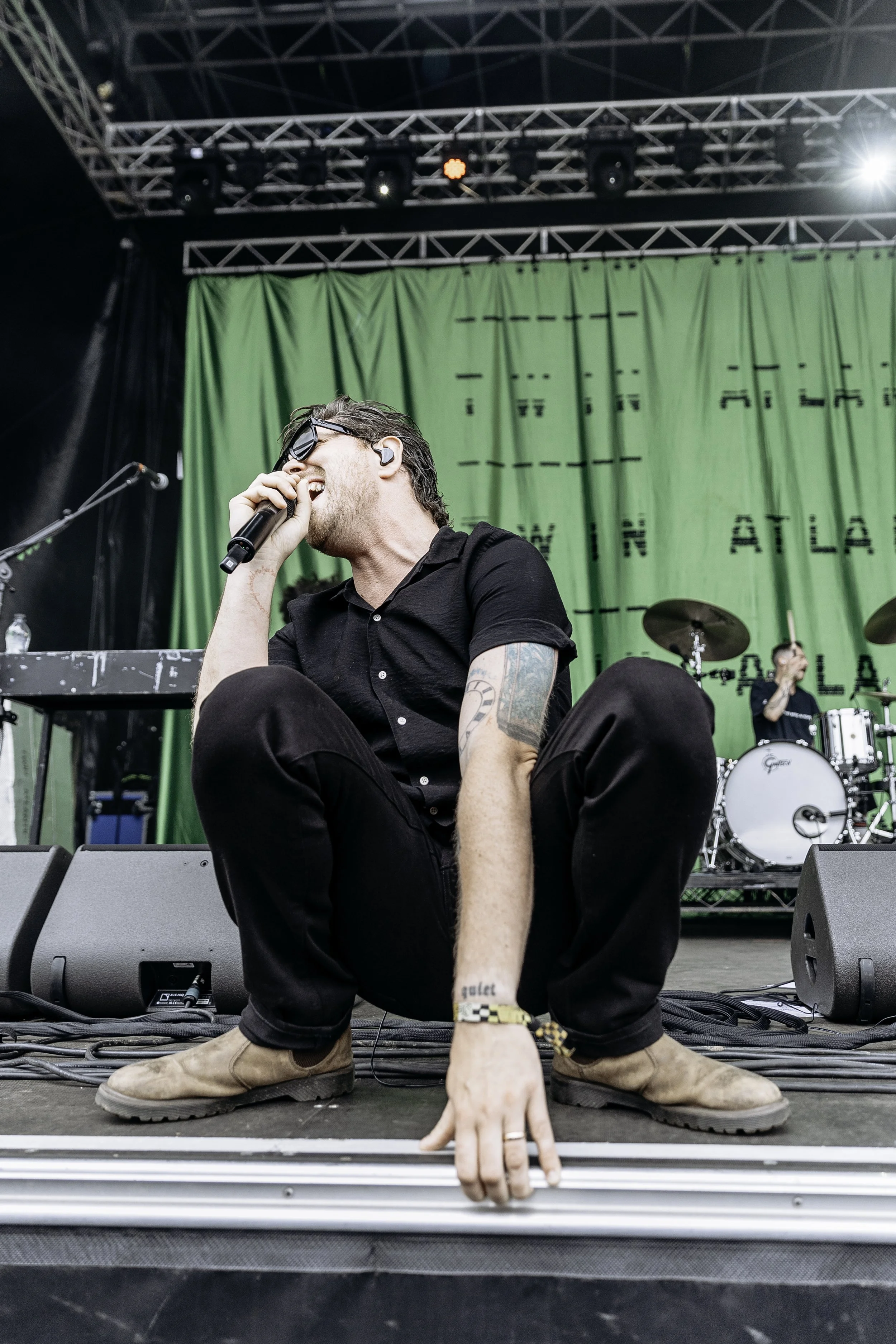 Male singer wearing sunglasses, black short-sleeve shirt, and brown boots, sitting on stage with green backdrop, singing into microphone, with drummer playing drums in background.