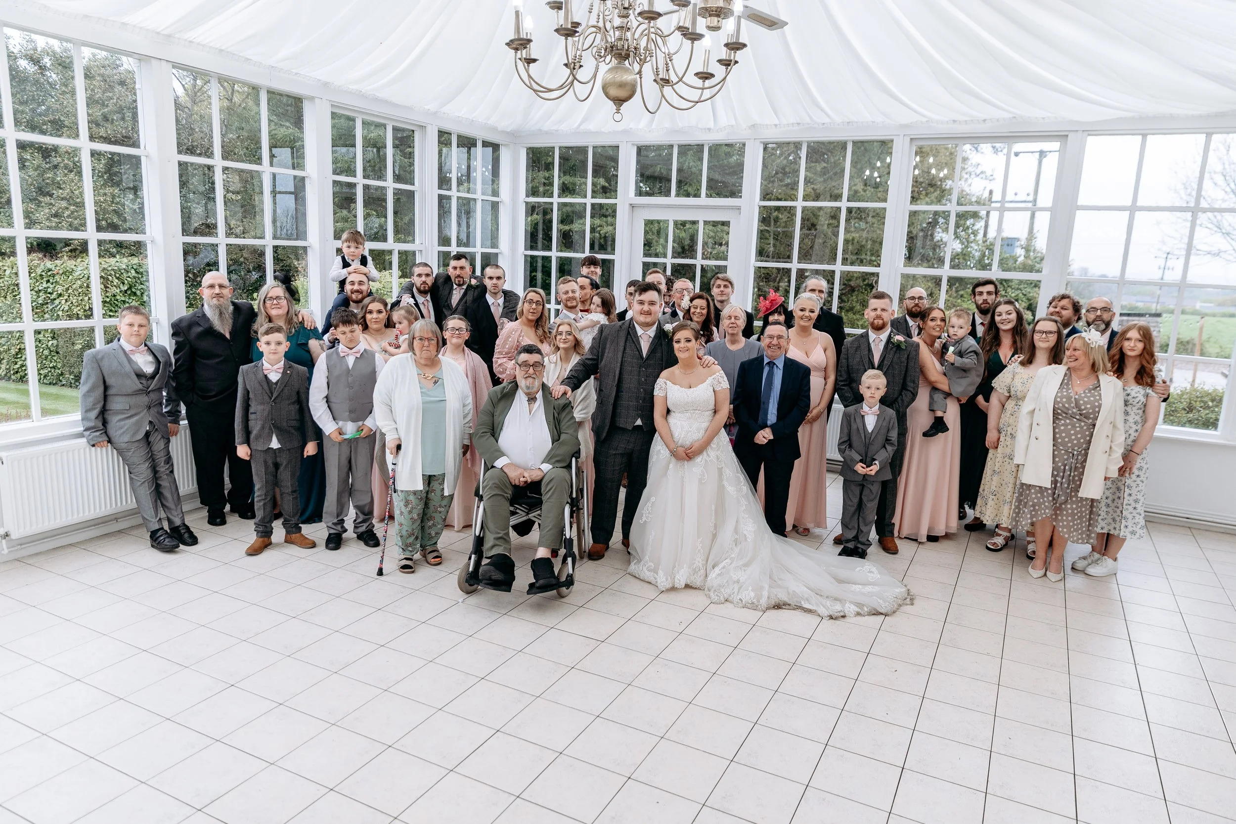 A large group of people in formal attire inside a white conservatory with large windows, gathered for a wedding celebration. The bride and groom are positioned at the front center, surrounded by family and friends, including children and elderly indi