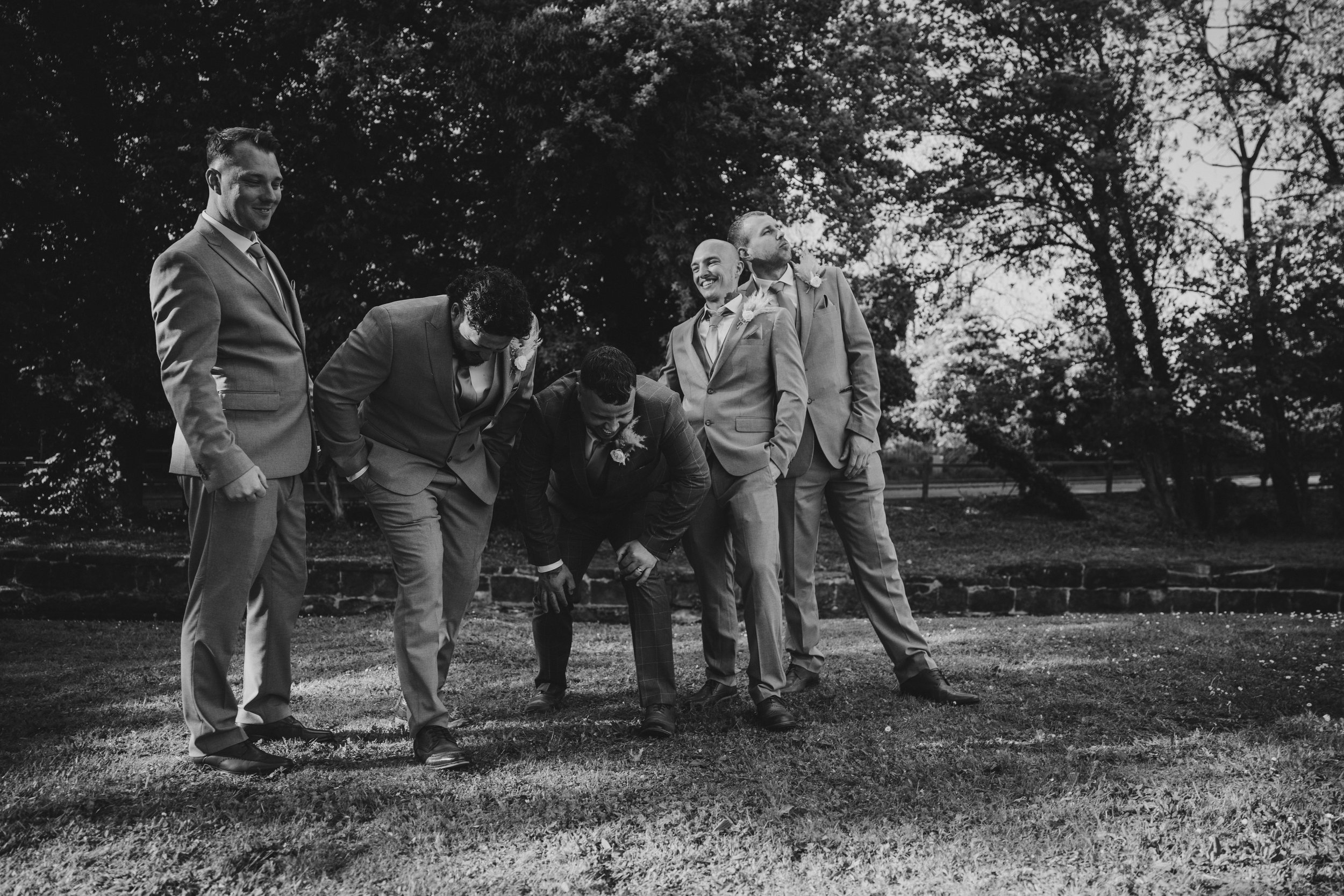 Black and white photo of five men in suits outdoors, posing playfully. One man is bending down while the others stand around him, some smiling and others looking up, in a park with trees in the background.