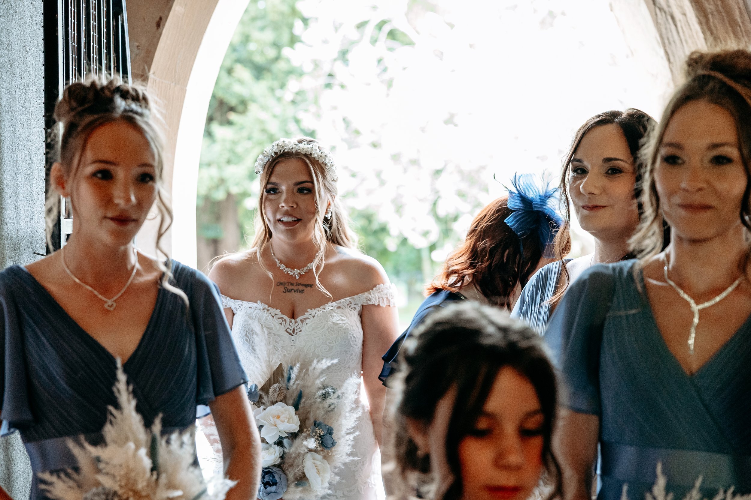 A group of women, including a bride in a white wedding dress with a floral crown, gathered in a hallway with an arched stone window and outdoor greenery in the background.