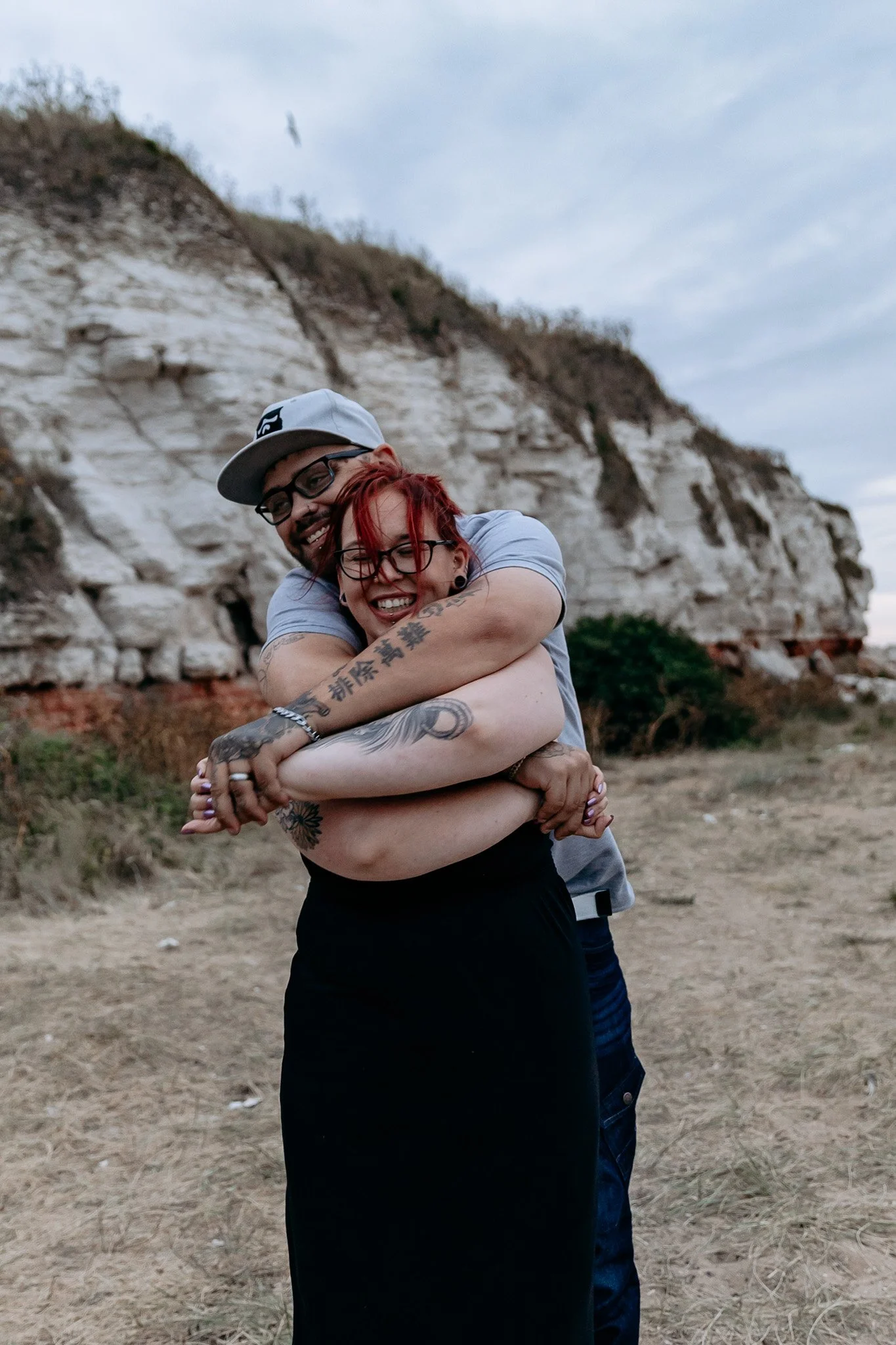 A man and woman hugging each other and smiling outdoors near rocky cliffs, with the man wearing a gray cap and glasses, and the woman with red hair and tattoos on her arms.
