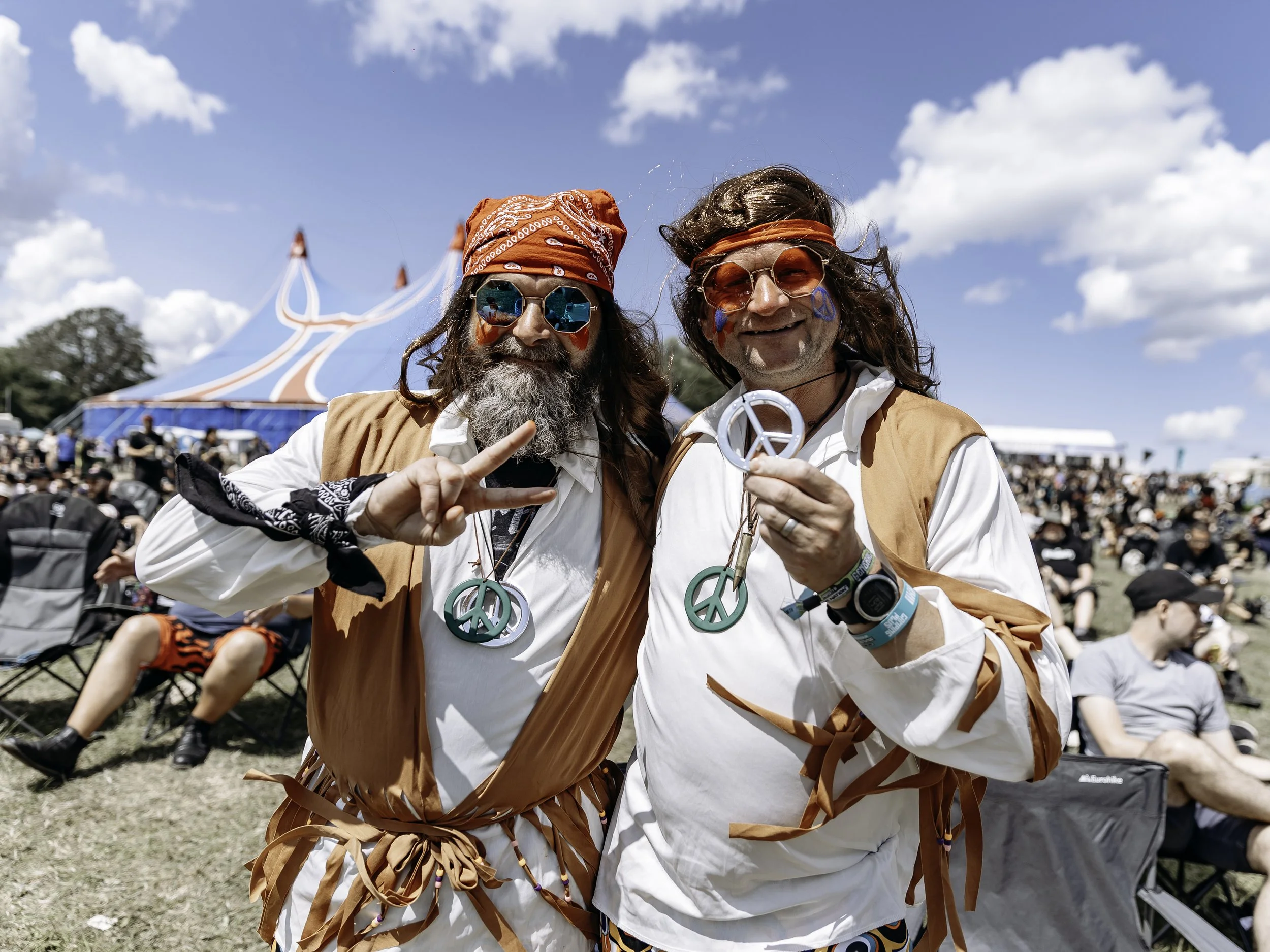 Two men dressed in 1960s hippie attire, wearing peace sign necklaces, sunglasses, and face paint, standing outdoors at a festival with a large crowd and a circus tent in the background.