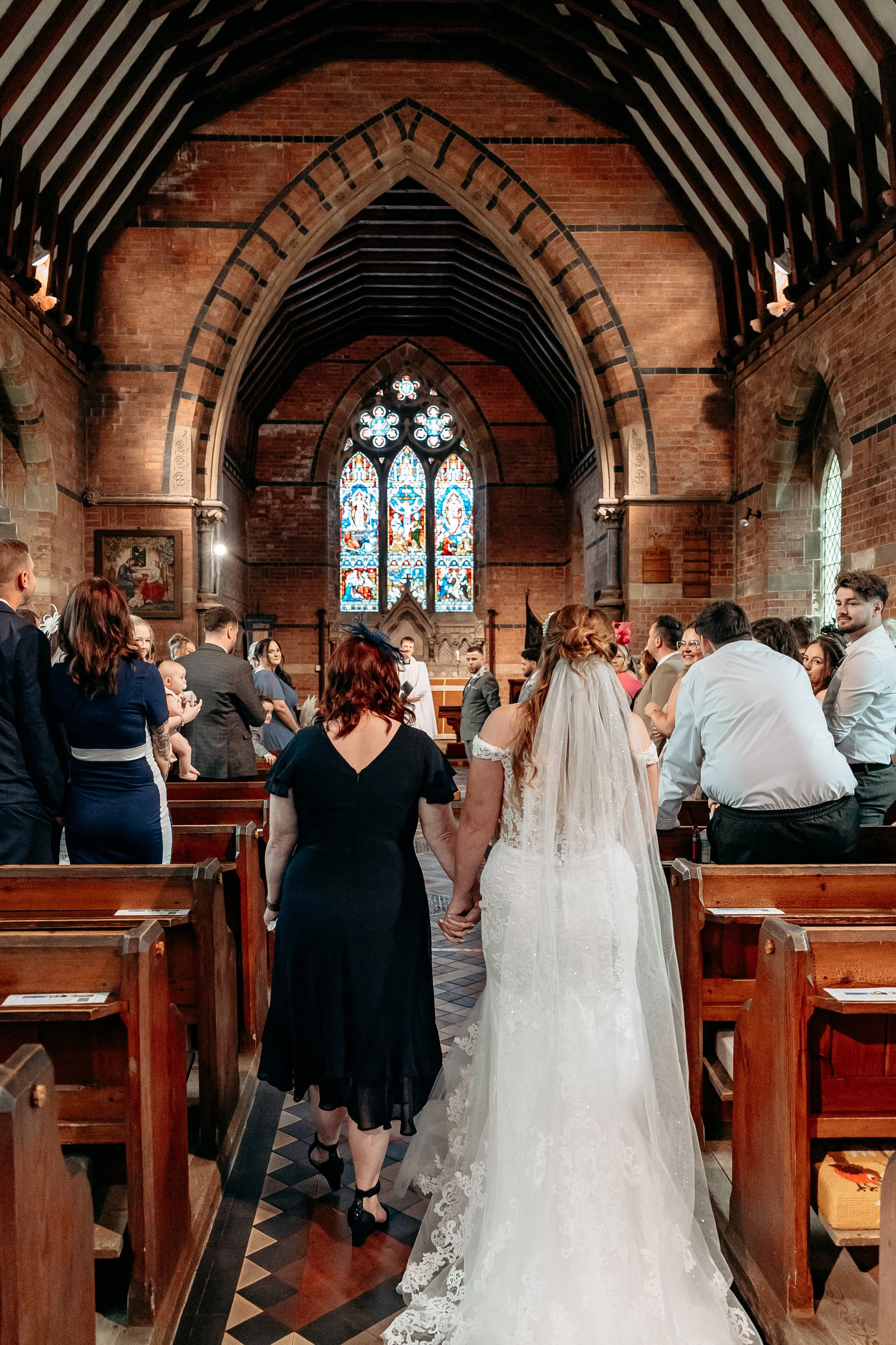 Bride and her partner walking down the aisle in a church during a wedding ceremony with guests seated on wooden pews and stained glass windows in the background.