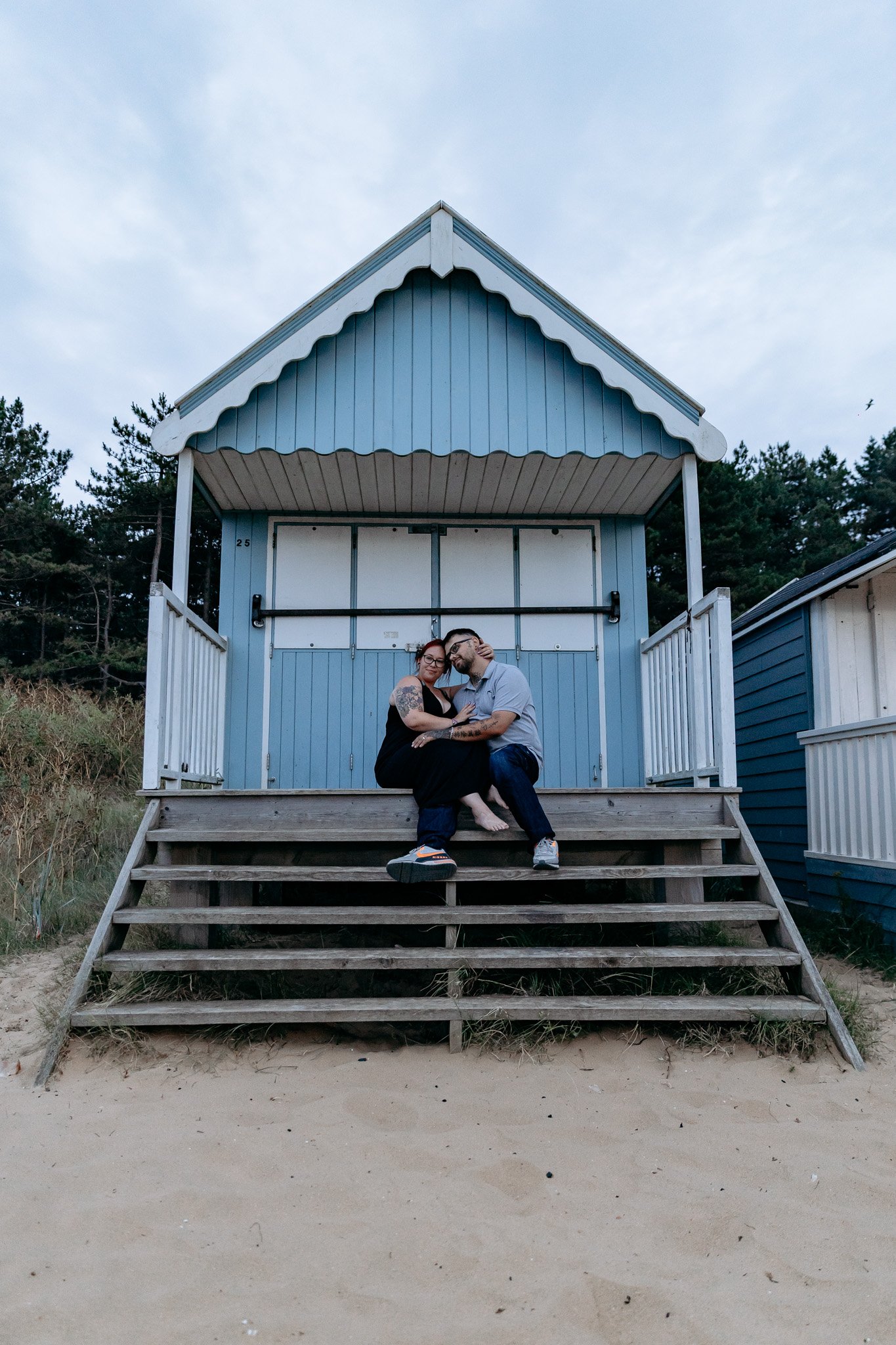 A couple sitting on steps of a blue beach hut, hugging and smiling, outdoors against a cloudy sky and trees.