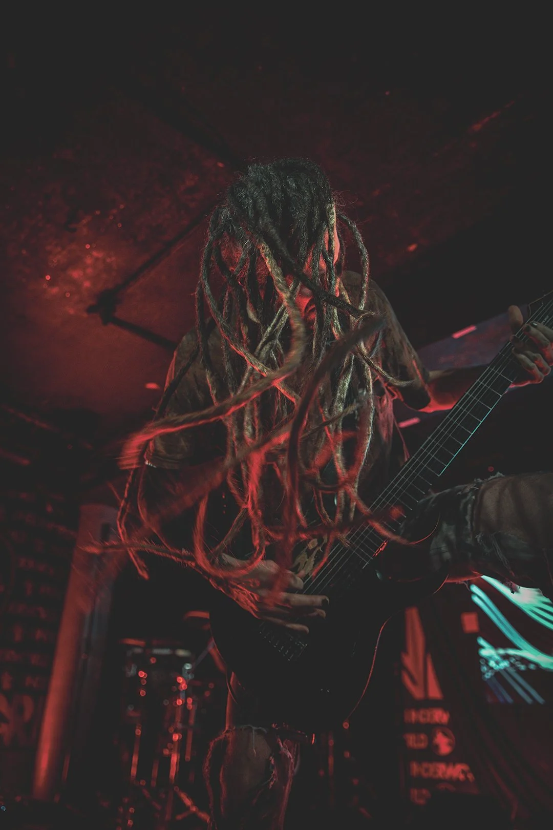 A musician with long dreadlocks playing an electric guitar on stage in a dark venue with red lighting.