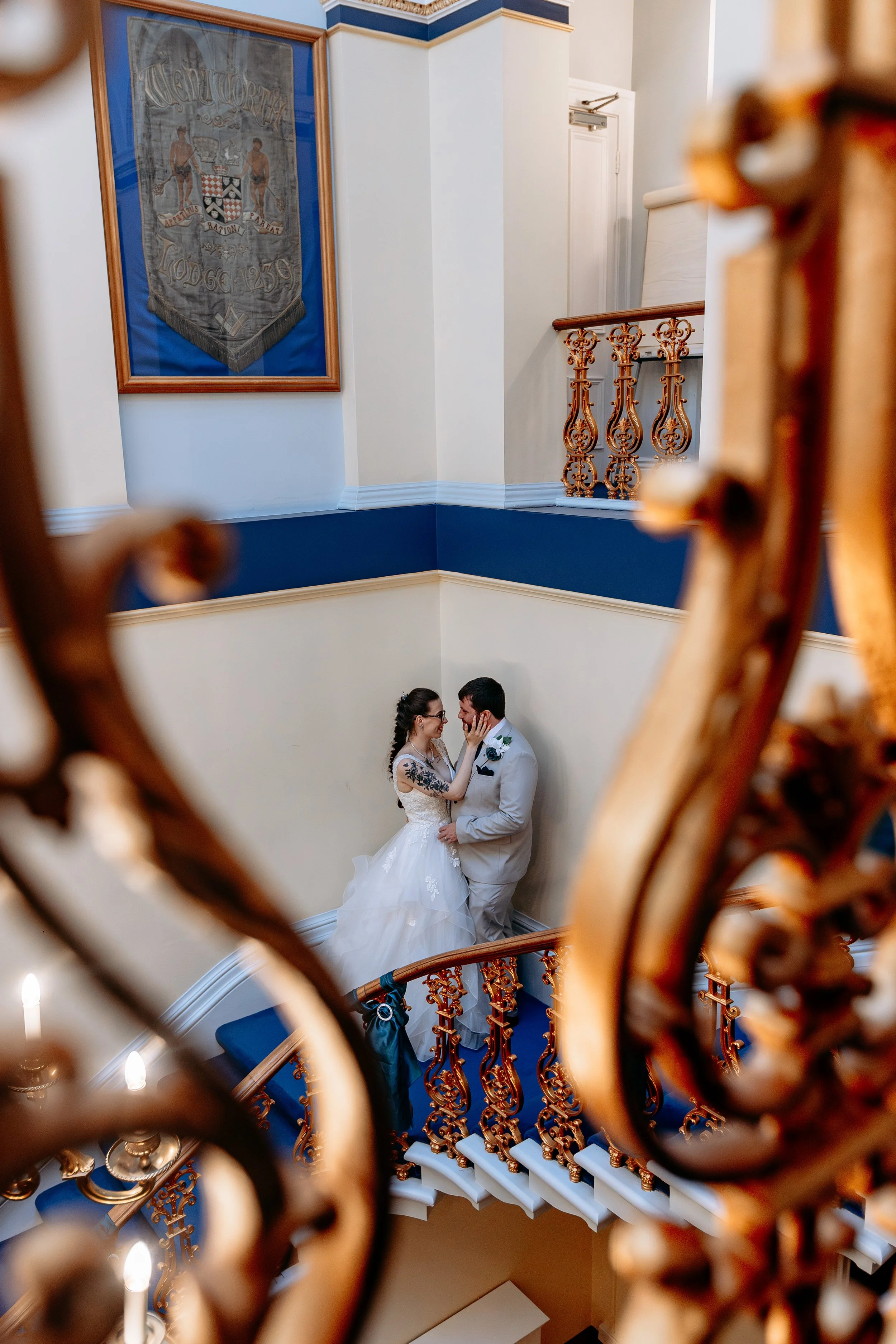 A bride and groom are standing on a staircase, touching foreheads and looking into each other's eyes, through a decorative wrought iron railing, with a framed vintage banner hanging on the wall behind them.