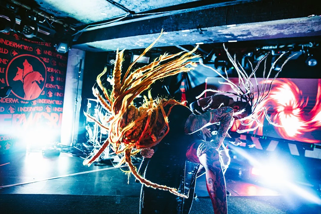 A performer with colorful dreadlocks on stage with bright lights and visual effects in the background.